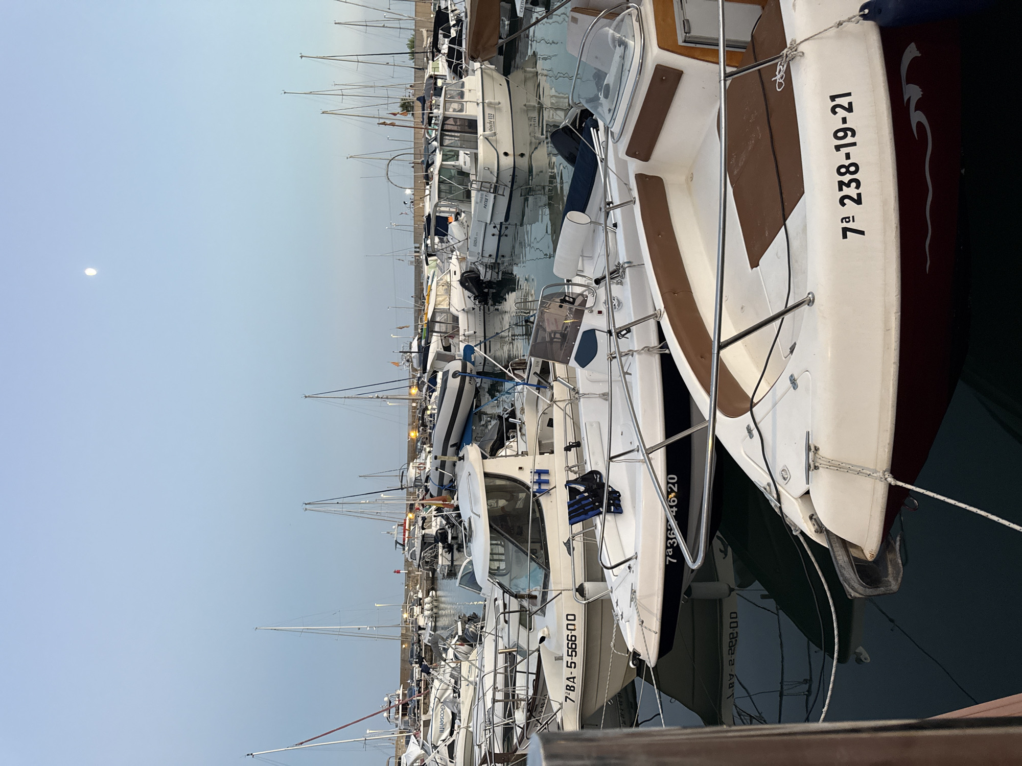 The image shows a marina in Blanes, Spain, likely at dusk or dawn, with a clear sky and a faint moon visible. Several boats are docked, ranging from small motorboats to larger yachts. The boats are predominantly white with some brown accents and are lined up neatly in the water. Some boats have registration numbers visible, such as "7ª BA-5 56600" and "7ª 362-46-20". The water is calm, reflecting the boats and the sky. The overall scene depicts a peaceful maritime setting.