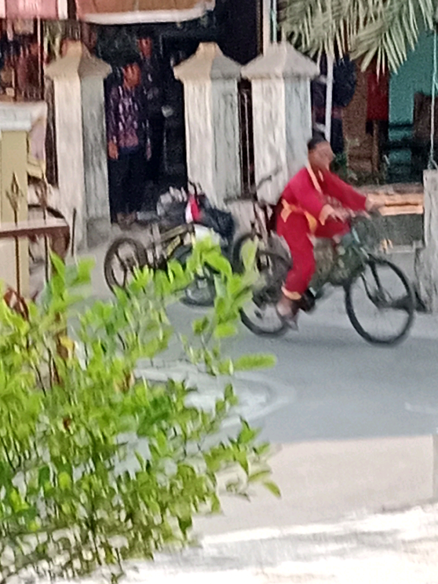 In Waru, Indonesia, a person in a bright red outfit is riding a bicycle. The bicycle is dark colored and appears to be a mountain bike. The rider's outfit consists of red pants and a red jacket with yellow trim. The person is wearing dark shoes. The background shows a street scene with architectural elements that suggest a commercial or residential area, including pillars and some foliage. To the left, there are some green leafy plants in the foreground, partially obscuring the view of the scene. There are also other people visible in the background, near some buildings. The lighting suggests it is daytime.