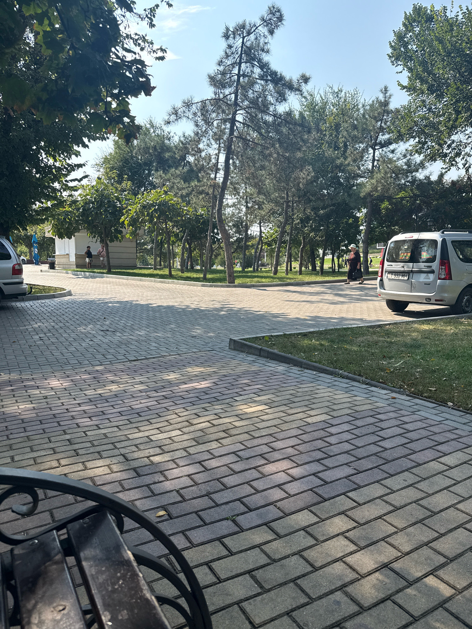 The image shows a park scene in Tiraspol, Moldova. In the foreground, a paved pathway made of grey rectangular bricks dominates the scene, with a dark metal bench visible on the left. Sunlight casts shadows across the pathway. In the middle ground, more paved pathways and green grass are visible. Several trees with green foliage line the pathways, and a small white building with a counter or window is situated amongst the trees. Two people, a child and an adult, are standing near the building. To the right, a silver minivan is parked on the grass verge next to the pathway. A woman wearing a dark dress and a light-colored hat is walking away from the minivan. The background consists of more trees and a clear blue sky.