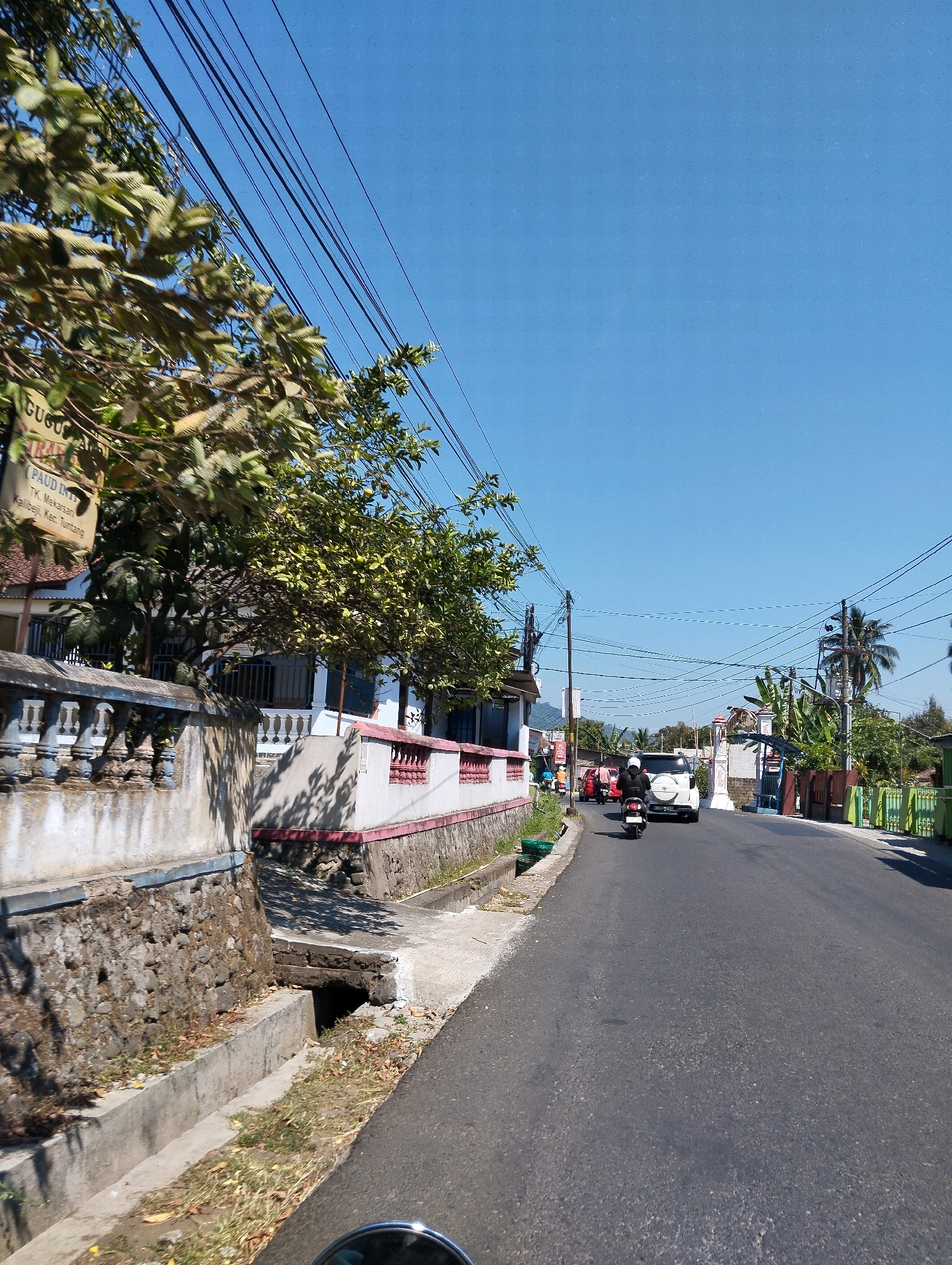 A low-angle, forward-facing, medium shot captures a sunny day on a paved road in Kalibeji, Indonesia. Overhead, a tangle of black electrical wires crosses the clear blue sky. To the left, a stone and concrete wall runs alongside the road, partially obscured by lush green trees with visible foliage. A sign, partially visible on the left side of the frame, appears to be a building marker.
The road itself is mostly empty, with a white SUV and two motorcycles traveling away from the camera. The motorcycle closest to the viewer has a rider wearing a black jacket and helmet. The road has a rough asphalt texture. On the right side of the road, a green fence and various buildings with trees and palm trees are visible. A concrete drainage ditch runs along the left side of the road, with some dry leaves scattered in it. The overall scene suggests a quiet, suburban or rural road.