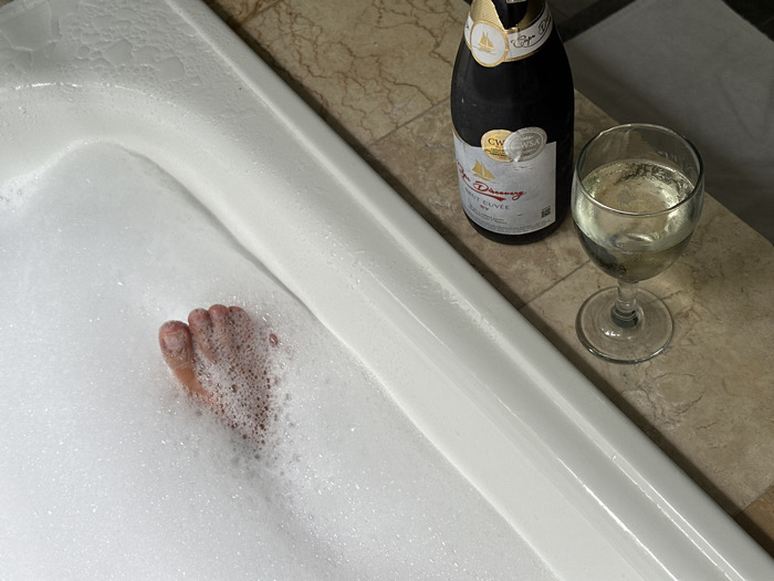 A white bathtub filled with bubbles has a foot partially submerged. Next to the tub is a bottle of Cape D'Aunay Brut Cuvee and a filled wine glass. The tub and bottle rest on a tan, tiled surface with a corner of a white towel visible in the background. The scene suggests relaxation and indulgence.