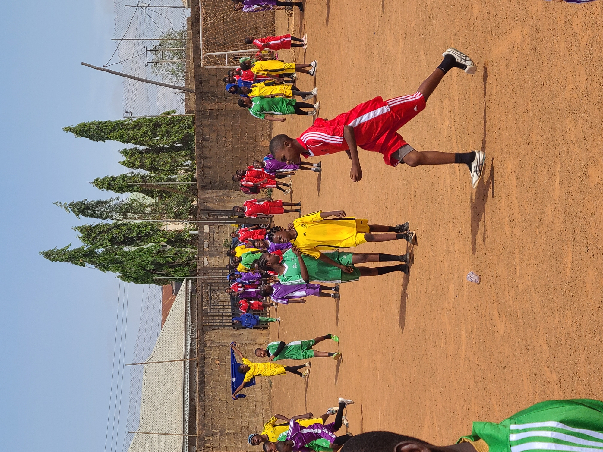 A wide shot captures a line of young soccer players, likely in Barakin Lerawi, Nigeria, performing warm-up exercises on a dirt field. The players, dressed in red, yellow, green, purple, and blue uniforms, are arranged in a row, stretching and raising their legs. A fence and some trees form a backdrop to the scene.