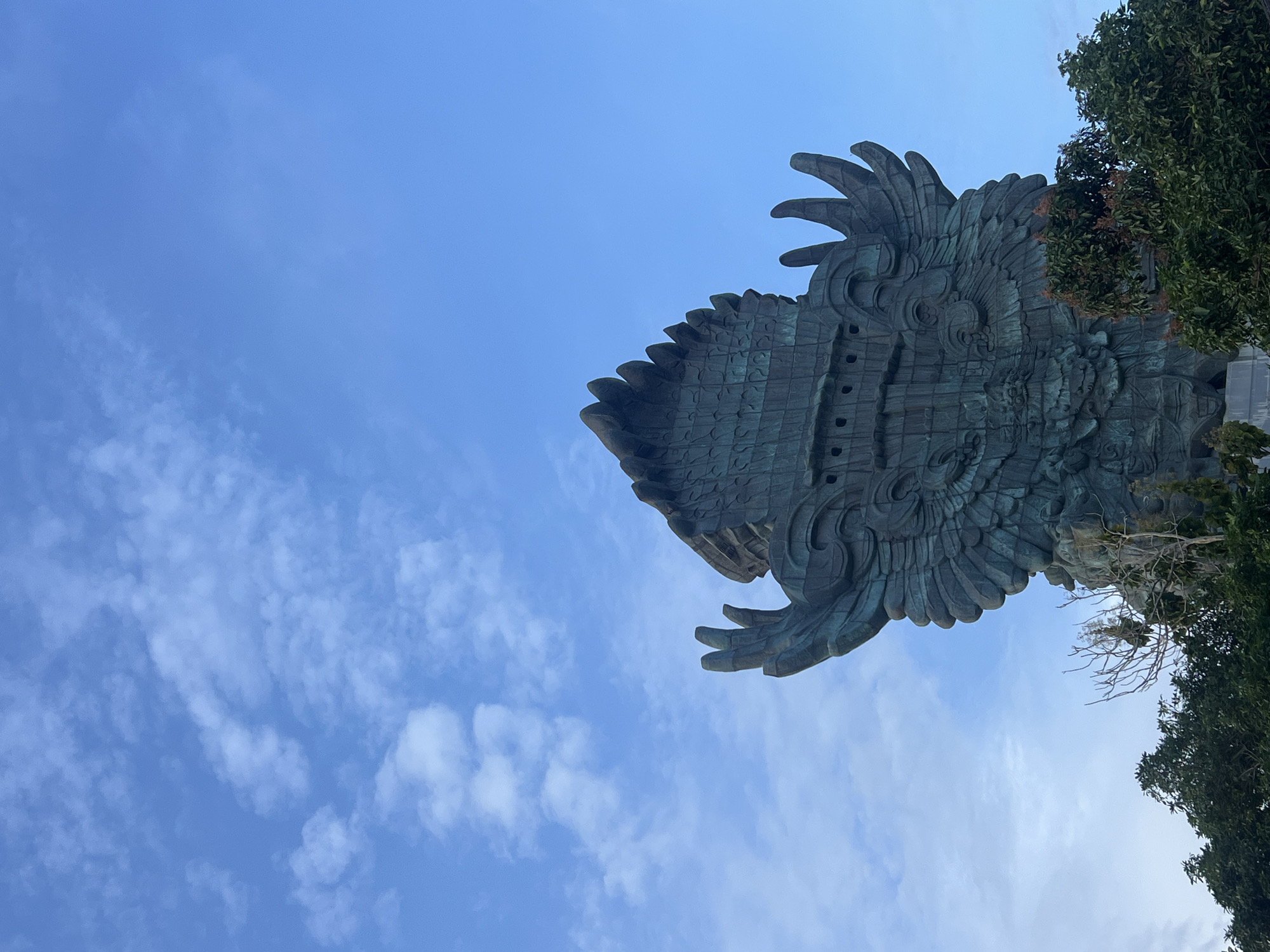 A large, intricately detailed segment of a colossal statue, likely depicting a mythical bird or creature such as Garuda, dominates the frame, set against a bright blue sky with scattered white clouds. The statue appears to be made of a dark metal, possibly bronze or copper, exhibiting a distinct green patina from weathering over time.
The visible section of the statue shows massive, stylized feathers and scaled patterns, suggesting the back or side of a wing and body. Intricate carvings and textures are visible across its surface, with some structural or decorative rectangular elements integrated into the design. The scale of the statue is impressive, as indicated by the low-angle perspective, looking up towards the monument.
The sky is a clear, vibrant blue, punctuated by soft, wispy white clouds, indicating fair weather during daylight hours. Green foliage from trees is visible along the right edge and bottom right corner of the image, partially framing the statue and suggesting an outdoor, landscaped setting. No human activity or specific events are depicted. There is no visible text in the image.
Given the provided context of Kuta Selatan, Indonesia, this is most likely a section of the iconic Garuda Wisnu Kencana (GWK) statue. The image is rotated 90 degrees clockwise from its likely intended upright orientation.