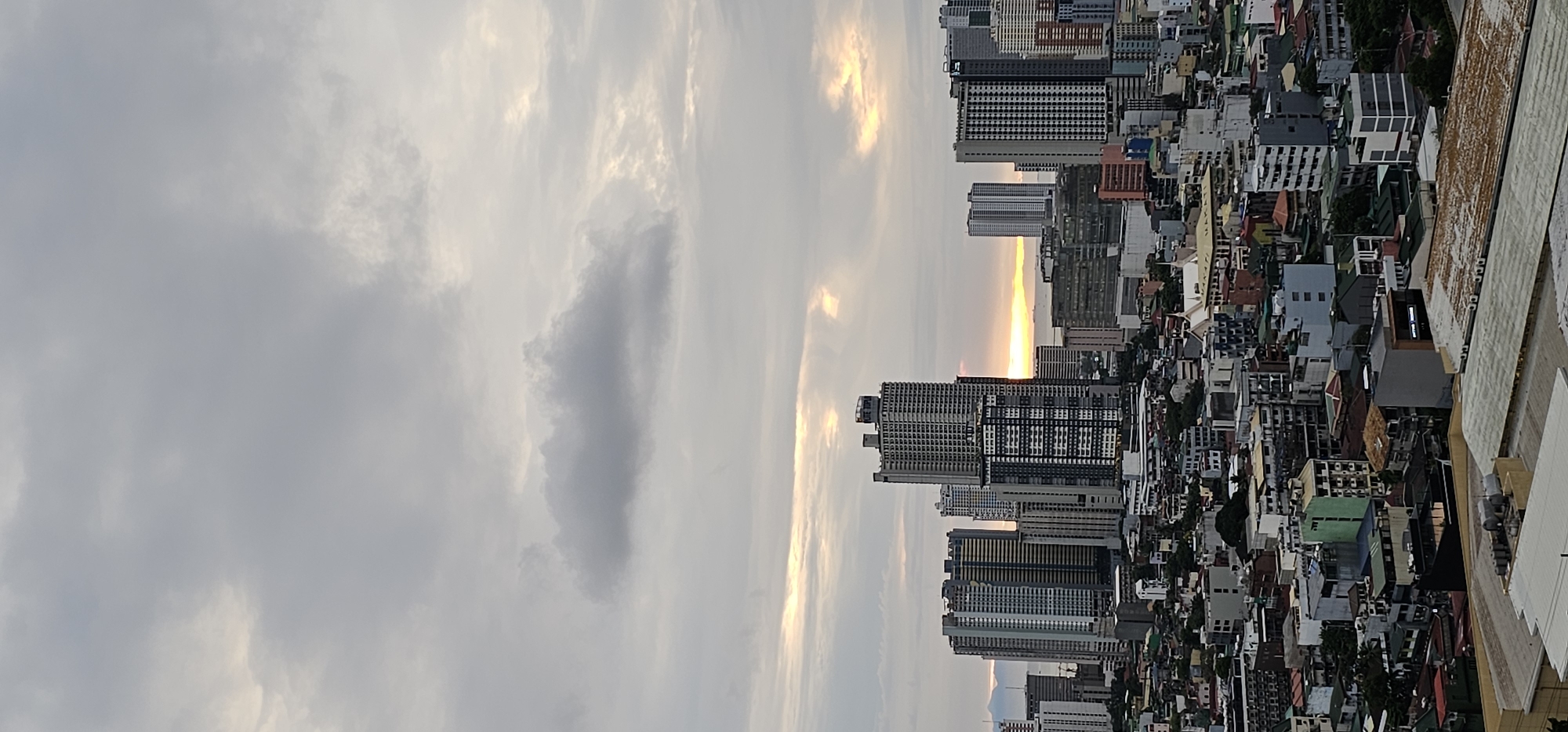 The image depicts an aerial view of a densely populated urban landscape, likely Makati, Philippines, characterized by a multitude of buildings of varying heights and architectural styles. The scene is dominated by skyscrapers and smaller residential or commercial structures, tightly packed together, creating a complex tapestry of urban development.
The time of day appears to be either dawn or dusk, as suggested by the soft, diffused light and the warm, golden hues visible in the sky and reflecting off some of the buildings. The sky is filled with clouds, some dark and dramatic, others wispy and luminous, indicating overcast or partly cloudy weather conditions. The overall mood is serene, with a hint of melancholy due to the muted colors.
In the foreground, a concrete structure, possibly a rooftop or balcony of a building, partially frames the scene, suggesting the vantage point of the photographer. The cityscape stretches towards the horizon, where it meets the sky. The buildings themselves display a range of colors, from neutral grays and whites to vibrant blues, greens, and reds on their roofs, adding a sense of dynamism to the otherwise monochromatic urban sprawl. Some of the taller buildings have grid-like facades, indicative of modern office towers.
No people are clearly discernible in the image, and the focus is on the built environment and the atmospheric conditions. There is no visible text within the image that provides specific information about the location or any ongoing events. The sheer density of buildings and the organized, yet chaotic, arrangement of the urban fabric are the most notable details, hinting at a bustling metropolis.