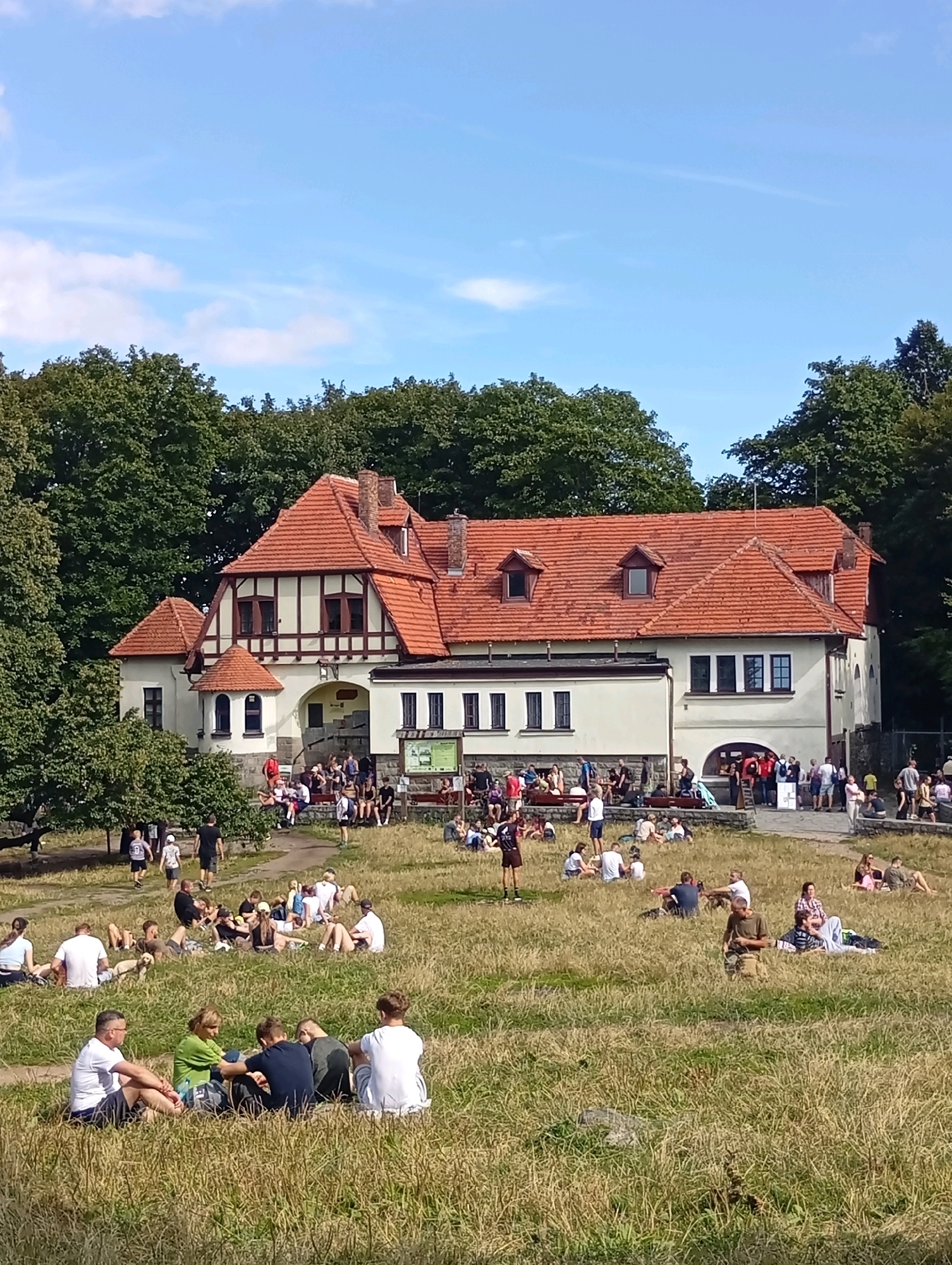 The image depicts a sunny outdoor scene near a building with red-tiled roofs, identified as the Sobótka observation post in Sobótka, Poland. A group of people are scattered across a grassy hillside in front of the building, some sitting and others standing. The building appears to be a historical structure, possibly a former military post, with a cream-colored facade and dark-framed windows. Trees line the background, suggesting a park or natural setting. The people are casually dressed, enjoying the warm weather. Some individuals are in small groups, while others are spread out individually, engaged in conversation or relaxation. The overall atmosphere is one of leisure and community gathering.