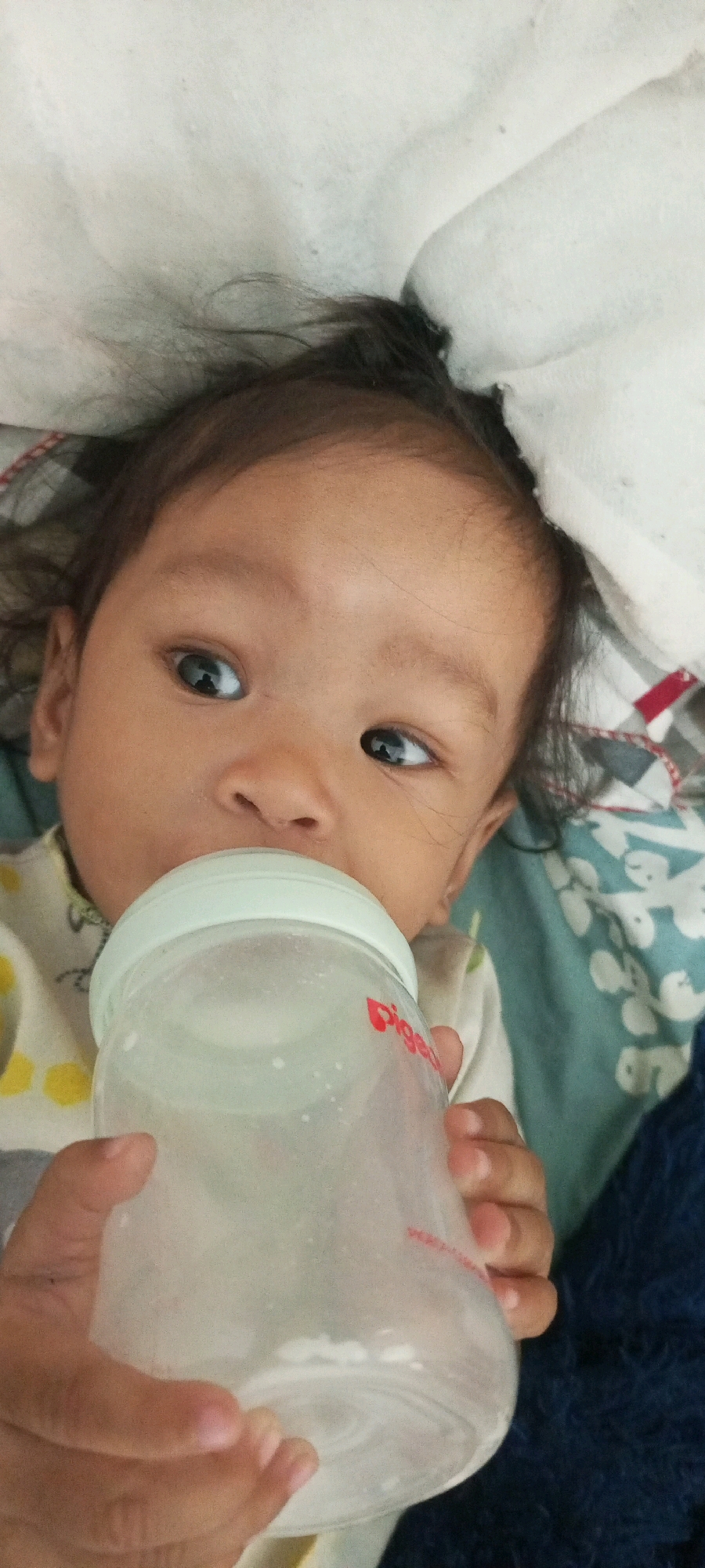 A close-up shot captures an infant being fed from a baby bottle. The baby, with dark hair and large, expressive eyes, is positioned near the top of the frame. The bottle, partially obscured by the baby's hands, is clear plastic with a white nipple and a faint red logo. The background suggests a domestic setting, possibly a bed, with a white pillow and a patterned blue and white blanket. The lighting is soft, indicating an indoor environment. The image appears to be taken in Sendawar, Indonesia, based on the prompt.