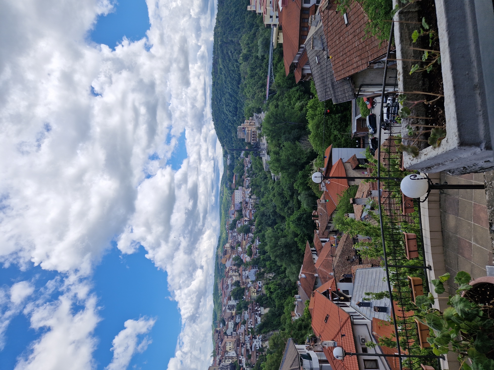 The image presents a scenic view of a town nestled at the foot of a lush, green mountain, under a sky filled with fluffy white clouds interspersed with patches of blue. The town itself appears to be densely populated, with buildings featuring terracotta roofs. In the foreground, there is a partially visible balcony, with flower pots and a railing indicating an elevated perspective.