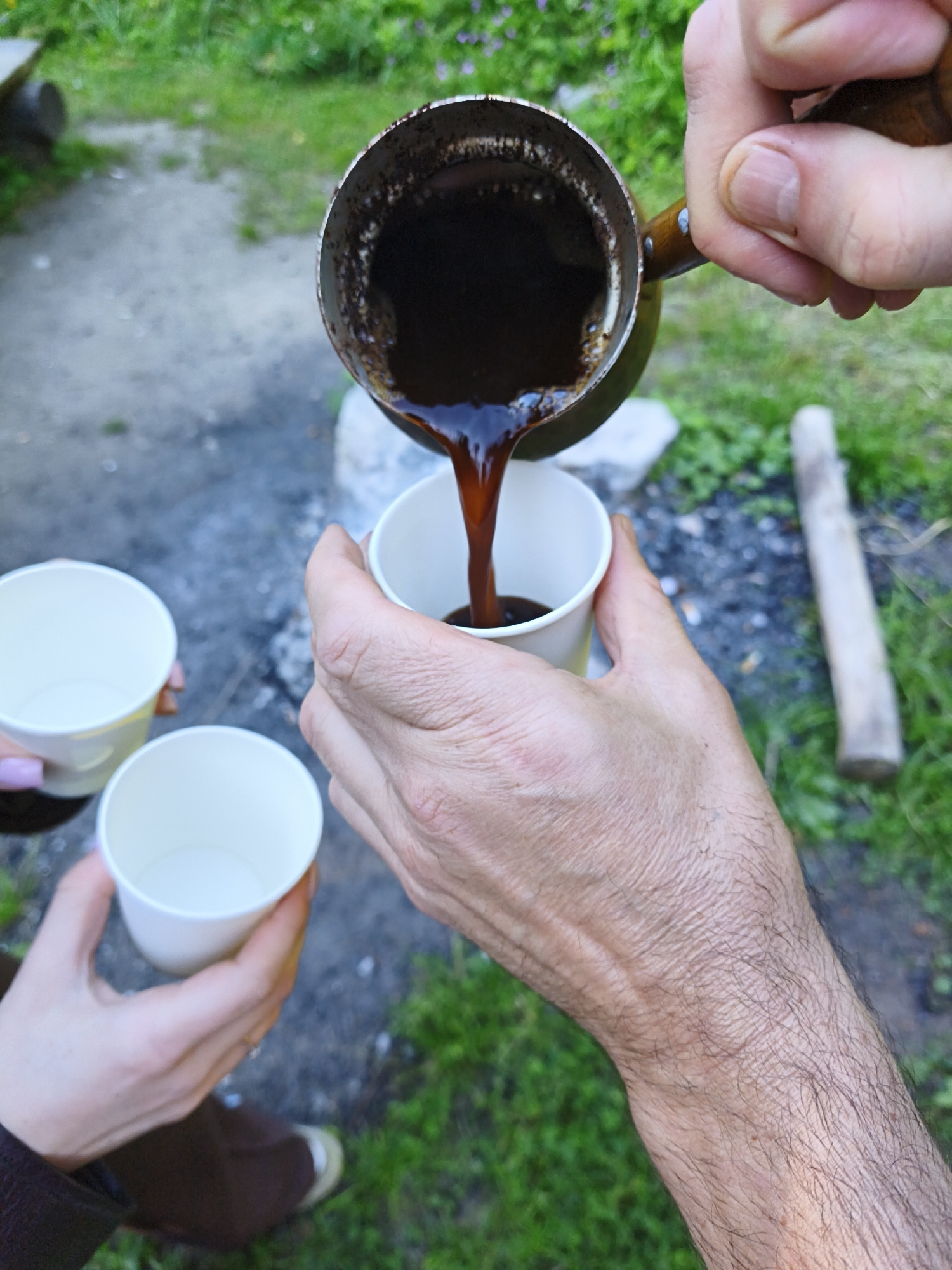 In Maardu, Estonia, two individuals are outdoors with green grass and a dirt path visible. One person's hand, with visible veins and dark hairs, is holding a metal cezve filled with dark liquid, pouring it into a white paper cup. Another hand, with light pink painted fingernails, is holding a separate white paper cup, positioned to receive the pouring liquid. A third white paper cup is also visible, held by someone whose hand has painted fingernails, but they are not actively participating in the pouring. In the background, a wooden object resembling a post or a piece of firewood lies horizontally on the ground amongst the grass. The scene suggests a casual outdoor gathering where coffee is being served.