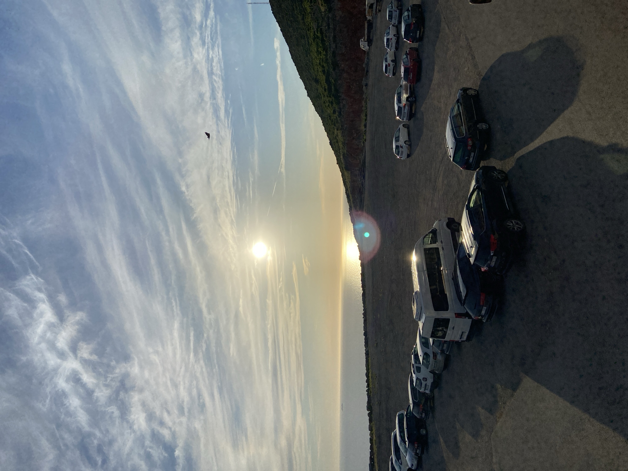 The image depicts a parking lot filled with various vehicles, including cars, a van, and possibly a small truck in the distance. The parking lot is situated on a gravelly surface adjacent to a body of water, likely the sea, which reflects the bright sun. A verdant, hilly landscape rises to the right of the parking lot, with a visible communication tower on its summit. The sky is partially clouded with wispy cirrus clouds, and the sun is visible near the horizon, casting a warm glow and long shadows across the scene. A single bird is in flight in the upper right portion of the sky. The location is identified as Kovačko Polje, Montenegro. There are no people visible in the image.