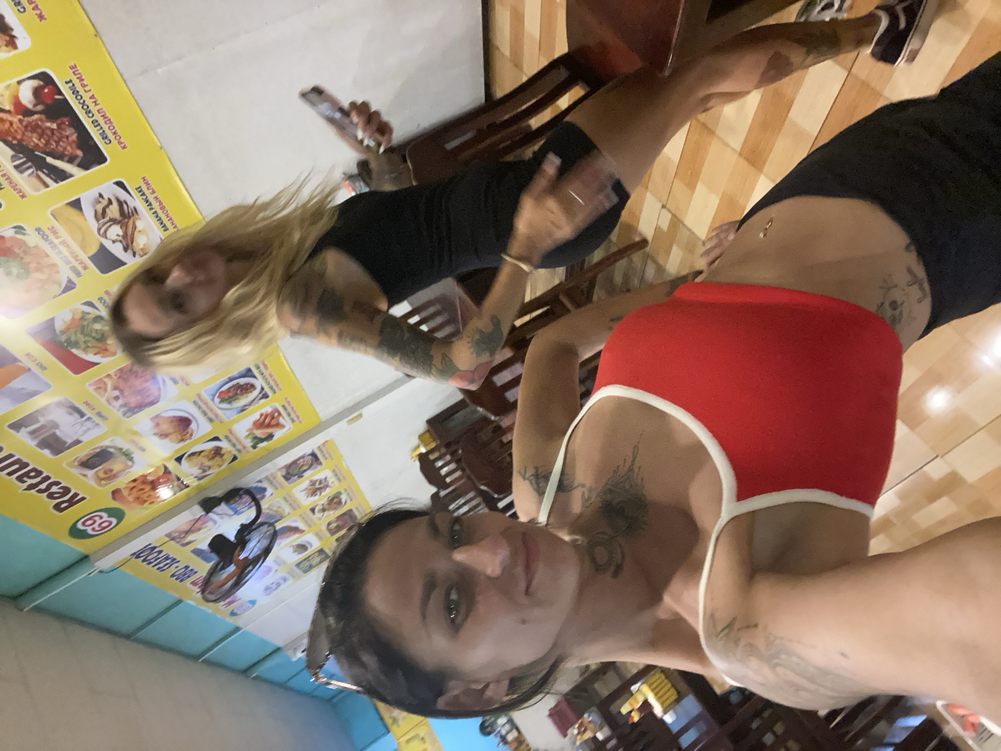 The media shows two young women in what appears to be a restaurant in Phan Thiết, Vietnam. The image is taken from a low angle, looking upwards. One woman, in the foreground, has dark hair and is wearing a red top. She has tattoos on her arms and torso. The second woman, in the background, has blonde hair and is wearing a black dress. She is holding a smartphone.
In the background, a menu is displayed on a yellow banner. Text on the banner includes "Restau", "BRO-SEAFOOT", "69", and various food items in English and Russian, such as "GRILLED CROCODILE", "BANANA PANCAKE", and "FRIED SEAFOOD". A ceiling fan is also visible. The setting seems to be indoors, with wooden tables and chairs. The lighting suggests it might be evening or indoors with artificial lighting. The overall mood is casual and appears to capture a moment of leisure.