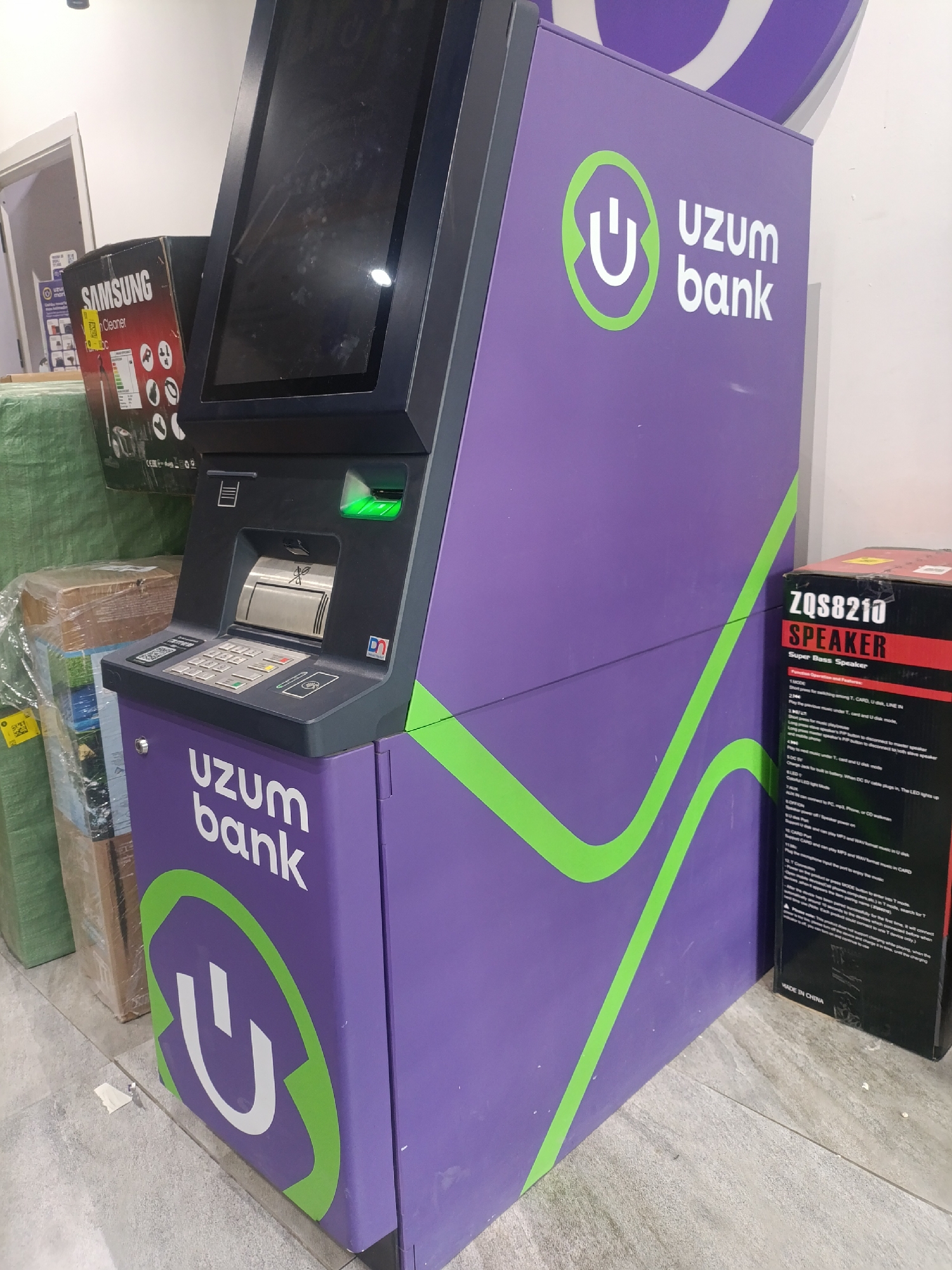 In the city of Karmana, Uzbekistan, a purple ATM with the "uzum bank" logo is the central subject. It is situated in a retail environment, evidenced by stacked boxes of merchandise to the left, including a Samsung cleaner box. To the right of the ATM, a black box labeled "ZQS8210 SPEAKER" is visible. The ATM features a touchscreen display, a keypad, a card slot, and a cash dispenser. The floor appears to be tiled. No people are present in the scene.