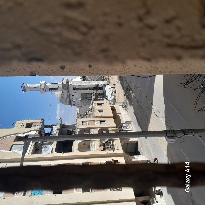 The image depicts a severely damaged urban street scene in Khan Yunis, Palestinian Territory, captured from an elevated perspective, likely a window or opening, on a clear, sunny day. The capture device is identified as a "Galaxy A14."
In the foreground, a textured, light-colored wall with two visible impact marks (consistent with bullet or shrapnel) partially frames the right side of the image, while a dark, out-of-focus vertical object frames the left.
The midground features a paved street, largely clear of vehicles, with substantial amounts of rubble and debris accumulated along its edges. A grey utility pole with numerous wires stands centrally on the left side of the street. Shadows cast across the road suggest mid-morning or mid-afternoon sun.
To the left of the street, a multi-story residential building, light-colored, exhibits extensive structural damage. Large sections of its facade are missing, exposing internal rooms, and windows are broken or absent. Some balconies are visible, one with fabric hanging. At the base of this building, on the far left of the street, two individuals are seated or crouched. Near the center of the street, a small figure in red clothing is on the ground.
In the background, beyond the damaged residential structures and street, a large mosque with a distinctive dome and a tall, slender minaret is visible. Both the dome and minaret show significant damage, including visible impact points and structural compromise. In the open area adjacent to the mosque, several makeshift shelters constructed from white tarpaulins are erected, and at least three small figures are discernible nearby. The sky above is clear blue with minimal cloud cover.