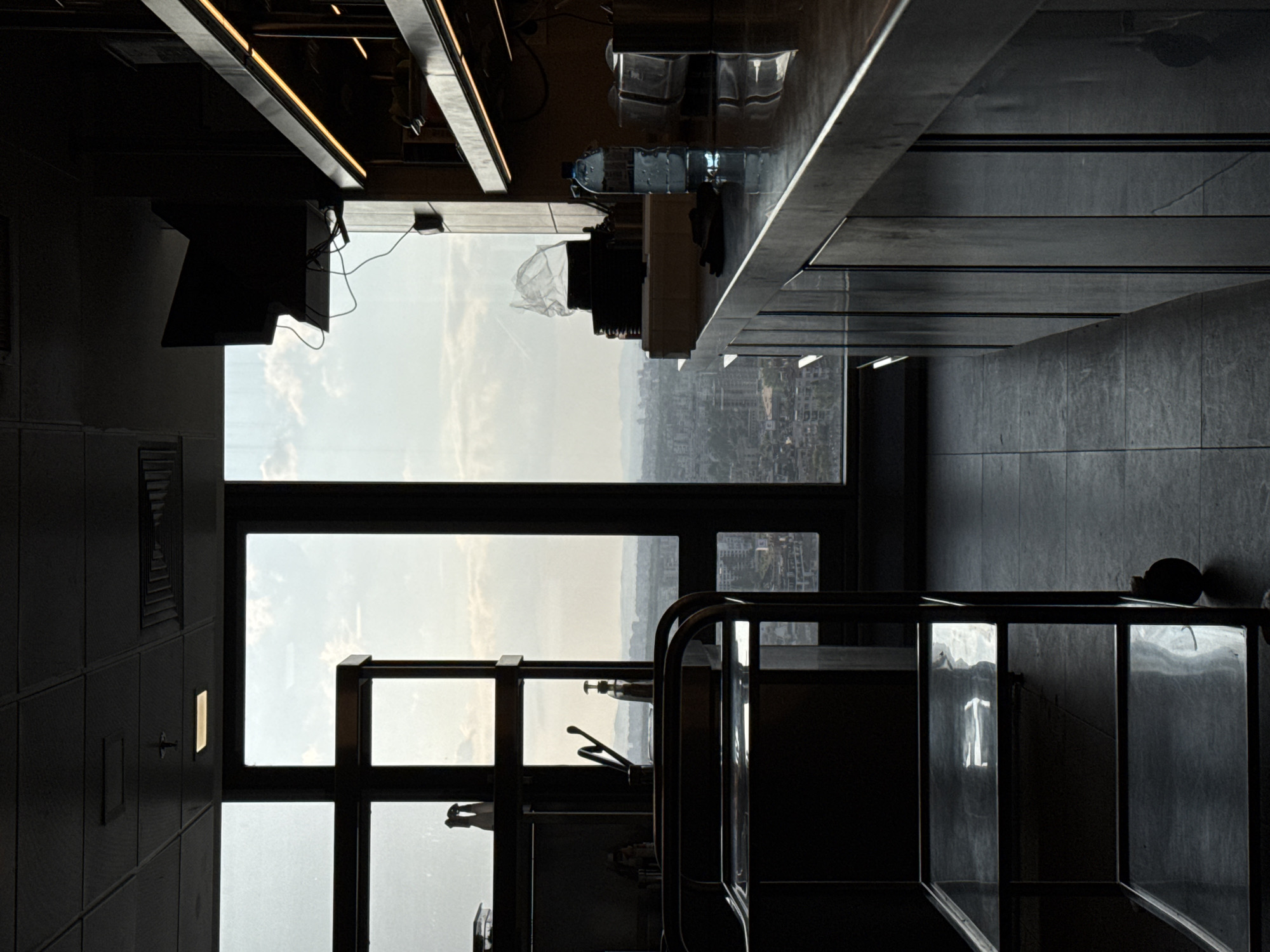 The image depicts a modern, minimalist kitchen or service area with a large window offering a panoramic view of a city, presumably Sofia, Bulgaria, in the distance. The interior is dimly lit, with the primary light source coming from the outside and some subtle artificial lighting above the countertops.
In the foreground, to the left, is a dark, metal utility cart with multiple glass shelves. The shelves are reflecting the light from the window, showing subtle distortions. To the right of the cart, a dark countertop runs along the wall, featuring a stainless steel sink, a bottle of water, and other unidentifiable objects. Above the countertop, illuminated shelves display a stack of dark plates and a plastic bag. A flat-screen television is mounted on the wall to the left of these shelves, angled slightly downwards, with visible wires.
The large window, divided by dark frames, dominates the background, revealing a sprawling cityscape with numerous buildings and distant mountains under a cloudy sky. The floor is tiled with dark square tiles. The overall atmosphere is one of quiet efficiency, possibly a professional kitchen, a bar, or a high-rise apartment with a view. No people are visible in the scene.