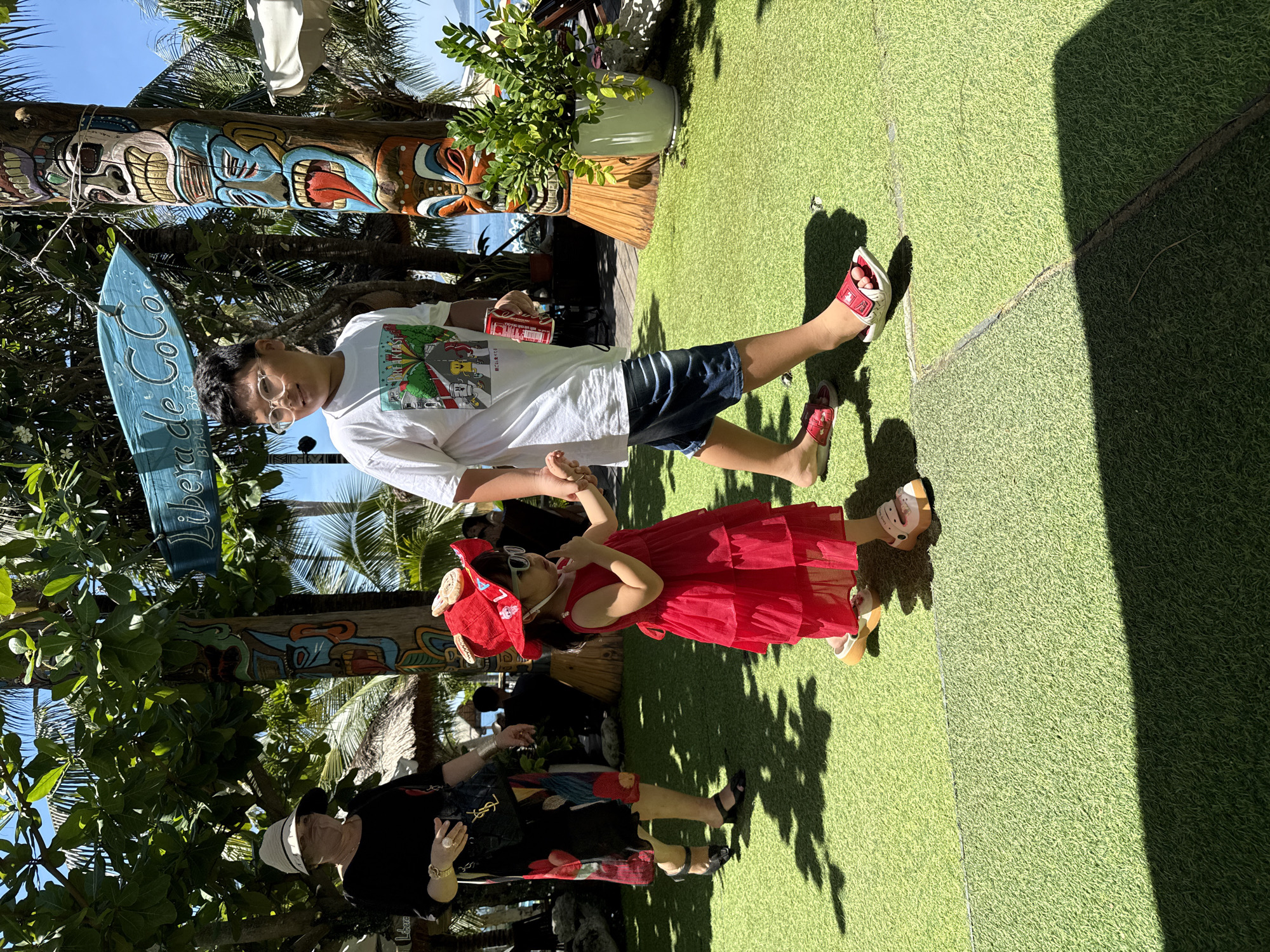 This is a vertical, full-body shot of two children and two adults standing on artificial turf. The lighting suggests it is daytime, with strong shadows cast by the sun. The setting appears to be a tropical resort or beach bar, with palm trees and tiki-style decorations visible in the background.
The subjects are:
- A young boy wearing glasses, a white t-shirt with a colorful graphic, and dark blue shorts. He is holding a red can.
- A young girl in a bright red, tiered dress and a red hat with a bow. She is wearing sunglasses and sandals.
- An adult woman wearing a black top with a colorful skirt, a white hat, and a gold watch.
- Another adult, mostly obscured by the girl, wearing dark clothing.
The children are positioned center-frame, with the boy on the left and the girl on the right, facing towards the right. The adults are in the foreground, on the left, with their backs mostly to the camera. The scene is set outdoors, with lush green foliage and a clear blue sky above.
Notable details include the vibrant colors of the tiki poles and the children's clothing. The artificial turf gives the ground a uniform green appearance. The overall atmosphere seems relaxed and summery, possibly a family outing or vacation.
Visible text on a blue surfboard-like object reads "Liber" and "de Coco BEACH BAR".