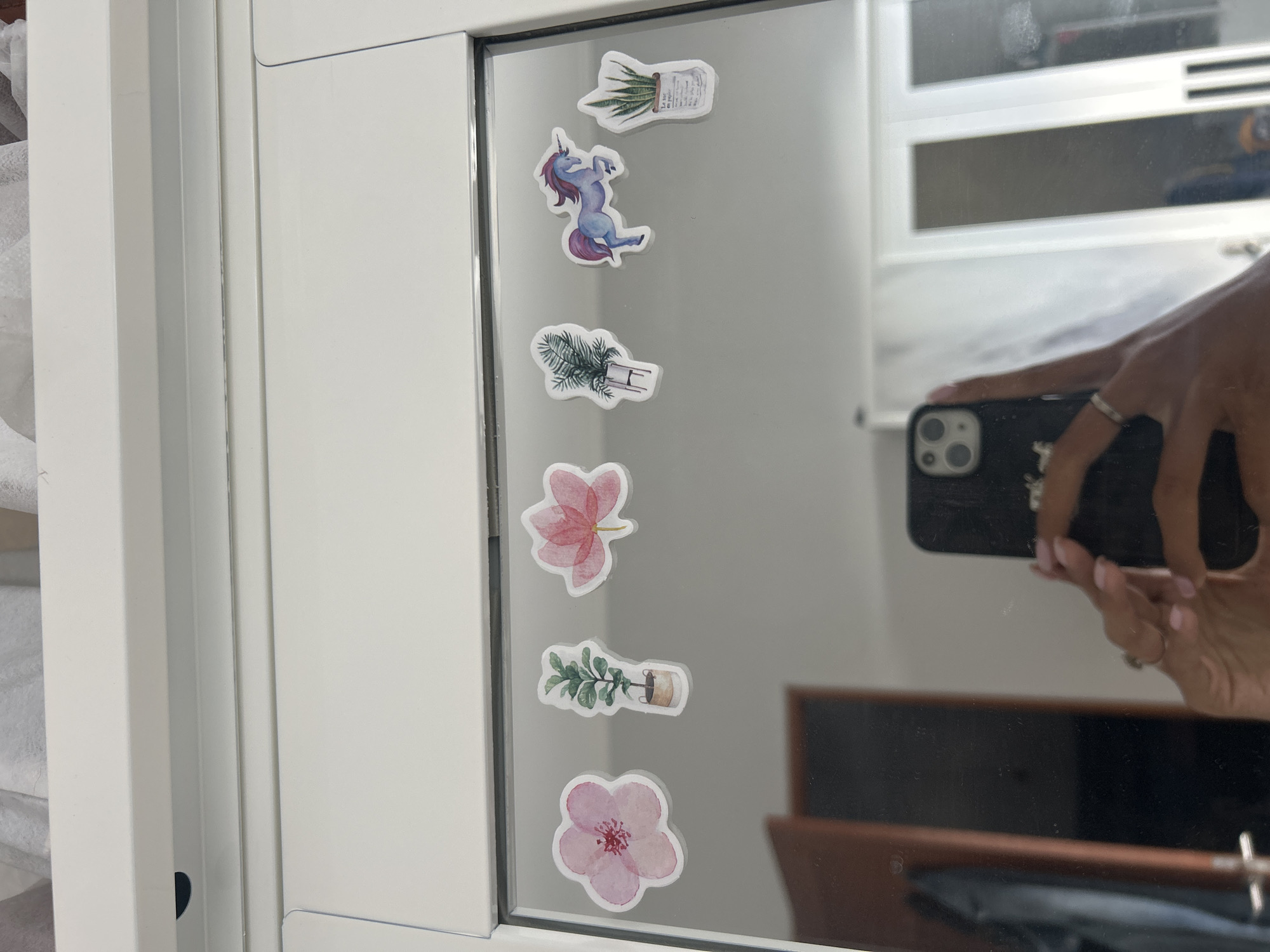 This image shows a close-up, eye-level view of a white mirrored closet door with several stickers affixed to its surface. The stickers, appearing to be watercolor illustrations, depict various items: a pink flower, a potted plant, a pink lotus flower, another potted plant with palm-like leaves, a blue and purple unicorn, and a potted snake plant in a paper bag. Below the stickers, the mirror reflects a person's hand holding a black smartphone, a doorway with a wooden door, and a portion of the room beyond, including what appears to be other closet doors. The stickers are arranged horizontally across the top portion of the mirror. The overall environment suggests an indoor setting, likely a bedroom or dressing area. There is no information available to link this image to Sukawati, Indonesia.