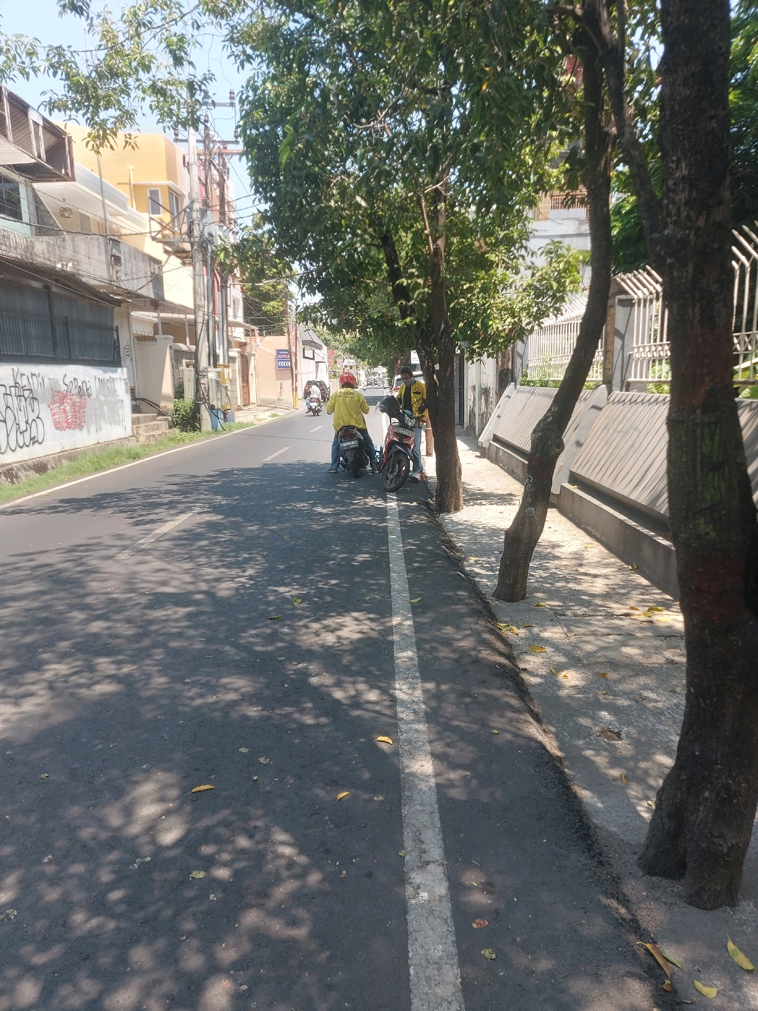 The image depicts a street scene in Makassar, Indonesia. The foreground shows an asphalt road with a white line marking the lane. Shadows from trees are cast across the road, and fallen leaves are scattered on the pavement and the adjacent sidewalk. On the right side of the road, a concrete wall with a decorative pattern runs along the sidewalk, bordered by trees. On the left side of the road, there are buildings with balconies and an exterior wall covered in graffiti. Several motorcycles are parked or in motion on the street. Two individuals are visible on motorcycles. One person, wearing a yellow jacket and a red helmet, is riding a motorcycle. Another person, wearing a mask and a yellow jacket, is also on a motorcycle, possibly accompanying the first. The sky is clear and blue, indicating a sunny day. The overall impression is a typical urban street with moderate traffic and pedestrian activity.