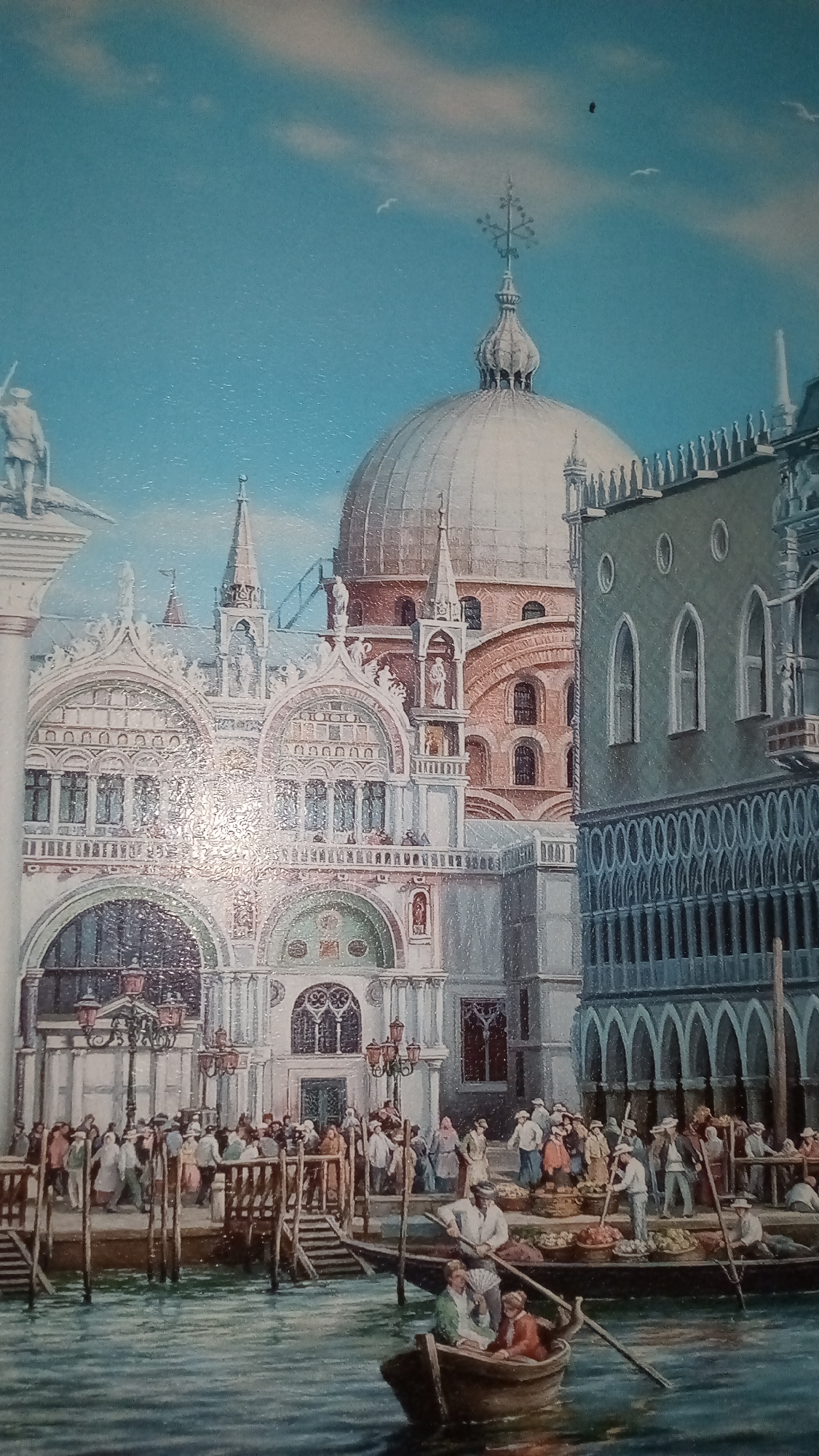 This is a painting depicting a bustling scene in Venice, Italy. The foreground features a canal with several gondolas. One gondola in the lower right has three people in it, one rowing and two passengers. Another gondola, larger and to the left, carries at least two people, one rowing. In the middle ground, a crowded boardwalk runs along the canal, lined with people engaged in various activities, some appearing to be merchants selling goods from baskets piled with produce. The background is dominated by grand Venetian architecture, including a large, ornate building with intricate stonework, arches, and statues, likely St. Mark's Basilica. To the right, a darker, more austere building with arched windows and a decorative facade stands. A large dome with a cross on top rises behind the basilica, suggesting it is part of a larger religious complex. The sky is a clear, bright blue with a few wispy clouds, and a couple of birds are visible. The overall impression is a lively, sunny day in Venice, with a vibrant atmosphere of commerce and daily life. The style of dress of the people suggests a historical setting, possibly the 18th or 19th century. There is no visible text in the image.