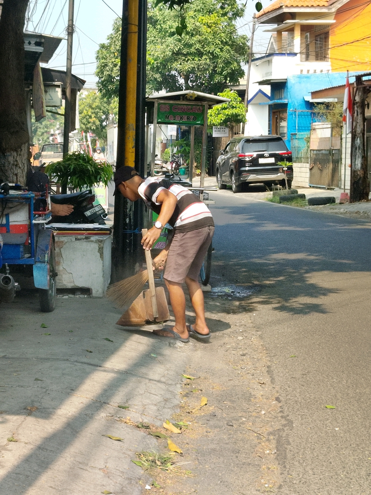 The image captures a street scene in Mojokerto, Indonesia. A man, wearing a black baseball cap, a striped t-shirt, and brown shorts, is sweeping the ground with a broom and dustpan. He is positioned on the side of a paved road, which appears to be in an urban or semi-urban area, indicated by the presence of buildings and parked vehicles.
To the left of the man is a three-wheeled vehicle, likely a type of local transport, painted blue and red, with a white container. A motorcycle is parked behind it. The background features buildings with various architectural styles and colors, including orange and blue. Green trees and foliage are visible, suggesting a generally warm climate.
A sign with text that appears to be in Indonesian ("ES CINCAU MOJOKERTO") is visible on a green structure in the mid-ground. The road surface is asphalt, and shadows from overhead structures and trees are cast across the scene, indicating a sunny day. The man's action of sweeping suggests a cleaning or maintenance activity. The overall environment appears to be a commercial or residential street.