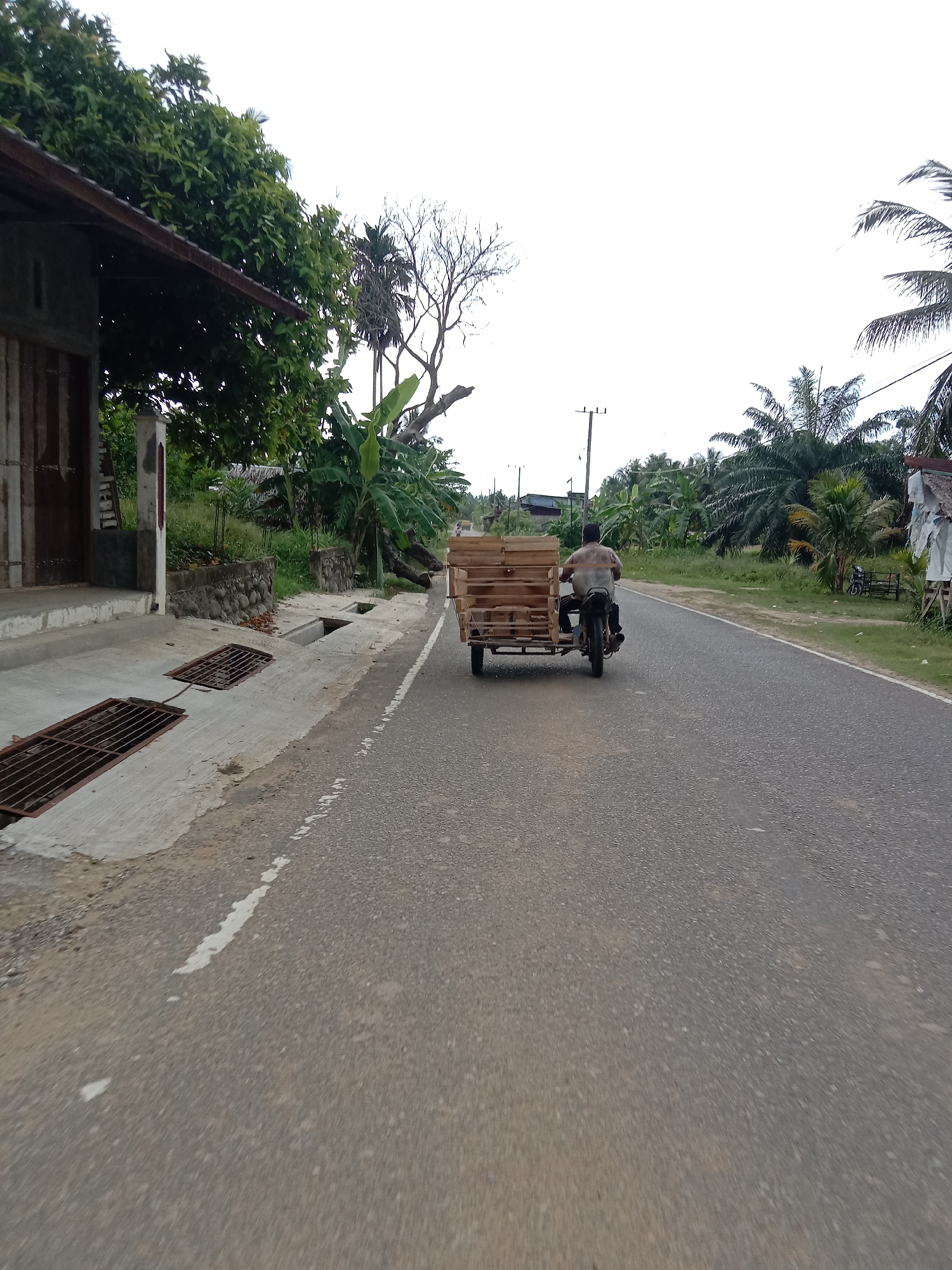 The image shows a road in Uleeblang, Indonesia. A man is driving a motorcycle with an attached sidecar, carrying a load of wooden crates. The road is asphalt and has white lane markings. Vegetation, including trees and banana plants, borders the road. A building is visible on the left side of the road. A motorcycle is parked on the right shoulder. The weather appears overcast.