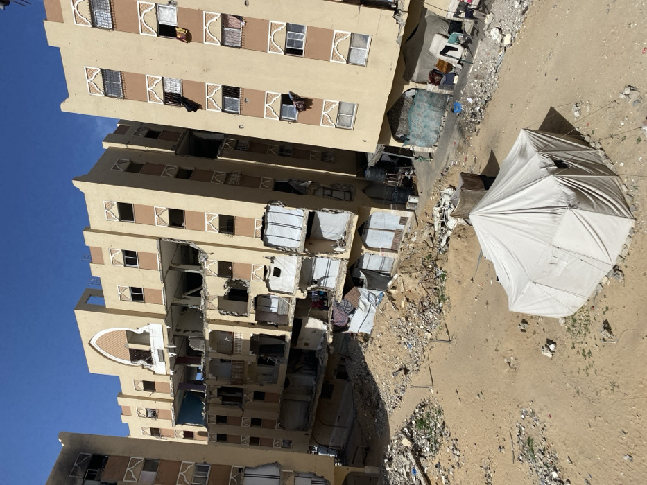 The image displays a severely damaged urban environment in Khan Yunis, Palestinian Territory, under a clear blue sky. The scene is dominated by multi-story residential or commercial buildings constructed of light-colored masonry, exhibiting extensive structural damage.
The central building features significant destruction across multiple floors, with large sections of exterior walls and balconies missing, exposing interior rooms and structural elements. Some areas show collapsed floor slabs and twisted reinforcement. An arched ornamental facade remains at the top center. To the left, another partially visible building also shows damage, including a blackened window opening on an upper floor. The building on the right, while appearing more intact structurally, has numerous broken or missing windows, some from which fabric items are visibly hanging. Makeshift coverings, possibly tarpaulins, are present in some ground-level openings of the damaged buildings.
The ground surface in the foreground and midground consists primarily of sand, heavily scattered with demolition debris, rubble, concrete fragments, and discarded refuse. A light-colored canvas tent, secured by ropes and rocks, is pitched in the sandy foreground, displaying visible windows on its sides. Smaller, darker, makeshift shelters fashioned from tarps are visible near the base of the buildings on the right. Plastic water tanks are also situated on stands near the damaged structures. No people are visible within the frame.