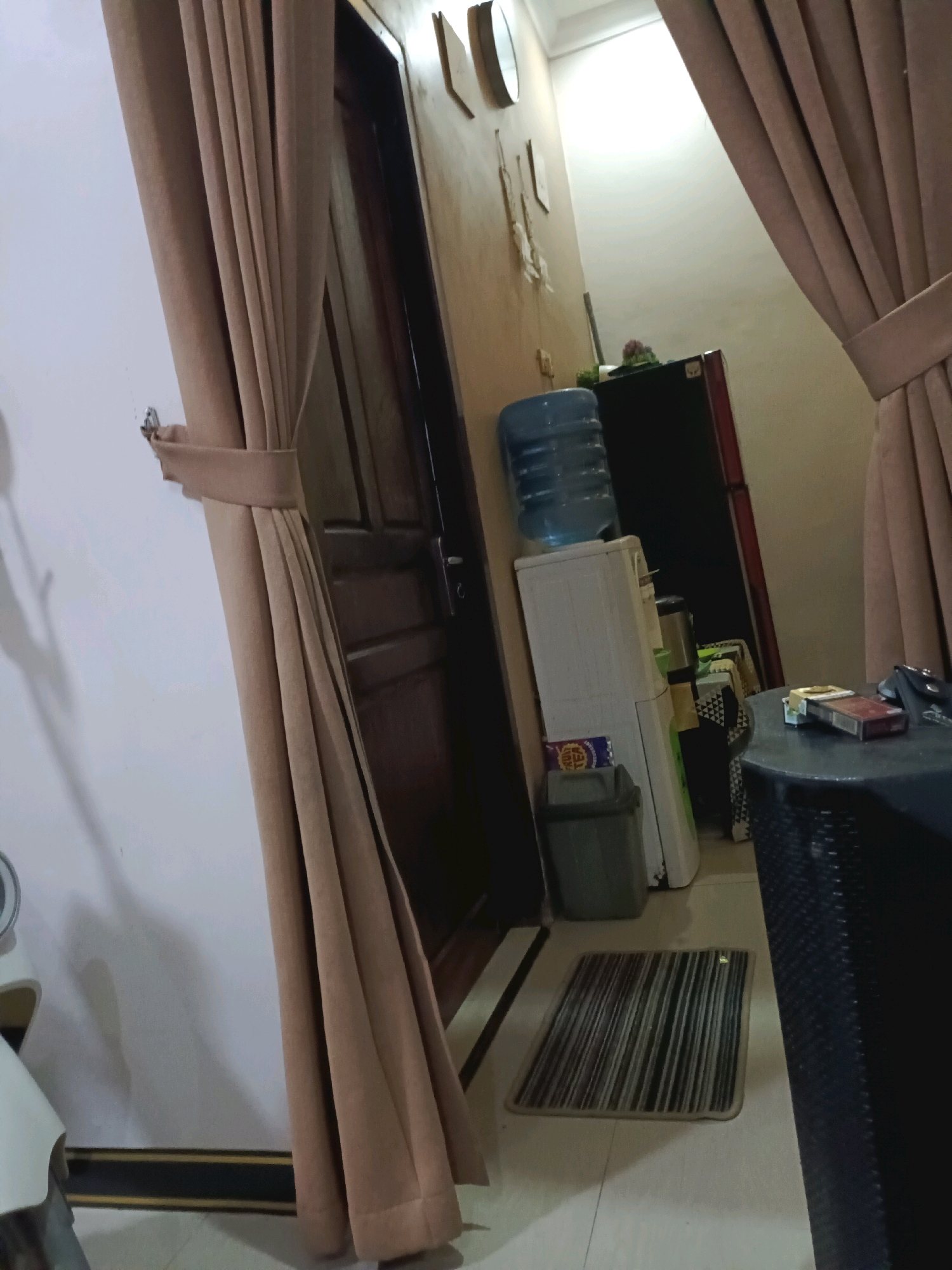 The image depicts an indoor scene that appears to be a hallway or a room within a residential dwelling in Rancang Kencono, Indonesia. The environment is lit, and the walls are painted a light color. On the left side of the image, a large, beige curtain is draped and tied back with a fabric band. Adjacent to the curtain is a white wall, with a shadow cast upon it. A brown wooden door is visible further down the hallway on the left.
In the center of the frame, a white water dispenser with a blue water bottle on top is situated. Next to the dispenser is a dark gray trash can containing some items. Behind the water dispenser, a dark refrigerator is partially visible. A small, patterned rug lies on the floor in front of the water dispenser.
To the right of the water dispenser and rug, a dark, circular table is present. On this table, a pack of cigarettes and a lighter are visible. Another dark object, possibly a basket or a piece of furniture, occupies the foreground on the right side of the image, partially obscuring the view of the table. A second beige curtain is also visible on the right, framing the scene. The floor is tiled, with some dark lines suggesting a patterned design. The overall impression is of a lived-in space.