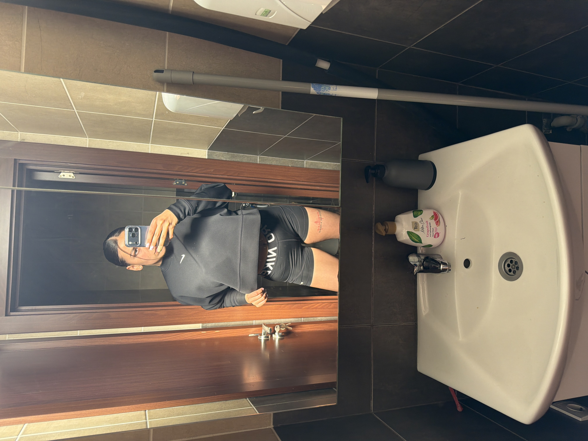 A mirror selfie is taken in a bathroom. Reflected in the mirror is a person wearing black Nike athletic wear, holding a phone taking the selfie. The bathroom has brown and beige tiles, a white sink with a bottle of soap on it, and a gray pipe along the wall. Keys are visible on a surface below the mirror. The location is Gabrovo, Bulgaria, based on the soap brand "Teo" which is a Bulgarian brand.