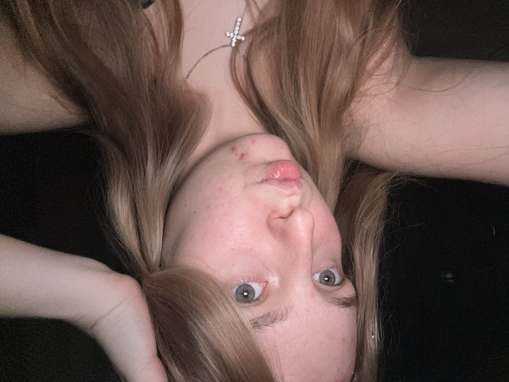 The image is a close-up, self-portrait of a young woman with blonde hair and light-colored eyes. She is looking directly at the camera with her lips pursed. She is wearing a necklace with a cross pendant. There are some blemishes on her chin. Her arms are raised and resting on her head. The background is dark and out of focus, suggesting a low-light environment. There is no indication of the city of Perm, Russia, or any other specific location in the image.
