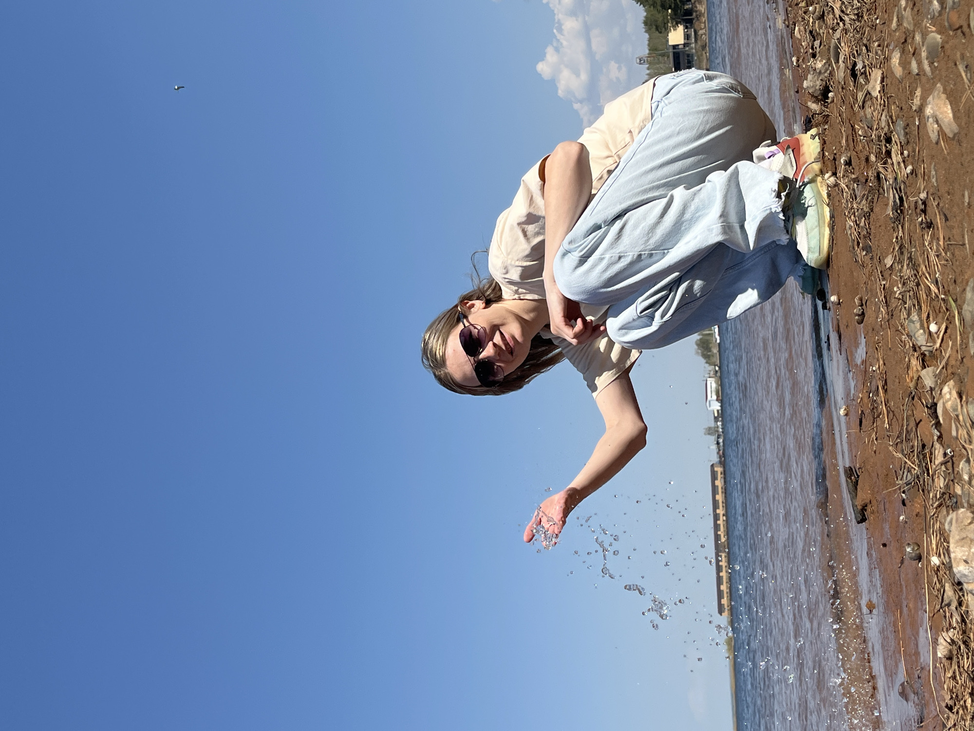 A person wearing a beige t-shirt, blue jeans, sunglasses, and colorful athletic shoes squats on the edge of a body of water, possibly a river or lake, and throws water with their right hand. The ground is sandy or muddy and littered with shells. In the background, structures resembling docks or buildings are visible on the opposite shore. The sky is blue with a few clouds. This photo was taken in the city of Chaikovsky, Russia.
