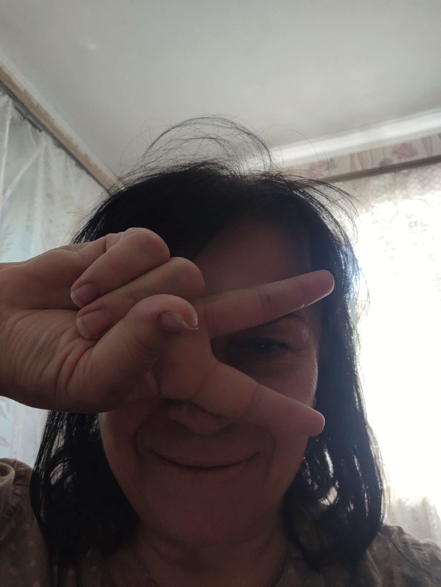 A woman is taking a selfie, holding her right hand up in front of her face in a peace sign gesture. Her dark hair is messy and falls around her shoulders. She is looking towards the camera with a slight smile. The background is blurred, but appears to be an indoor setting with light curtains and a patterned wallpaper. The lighting suggests it is daytime, with natural light coming from a window. The woman is wearing a light-colored shirt with a pattern of small dots.