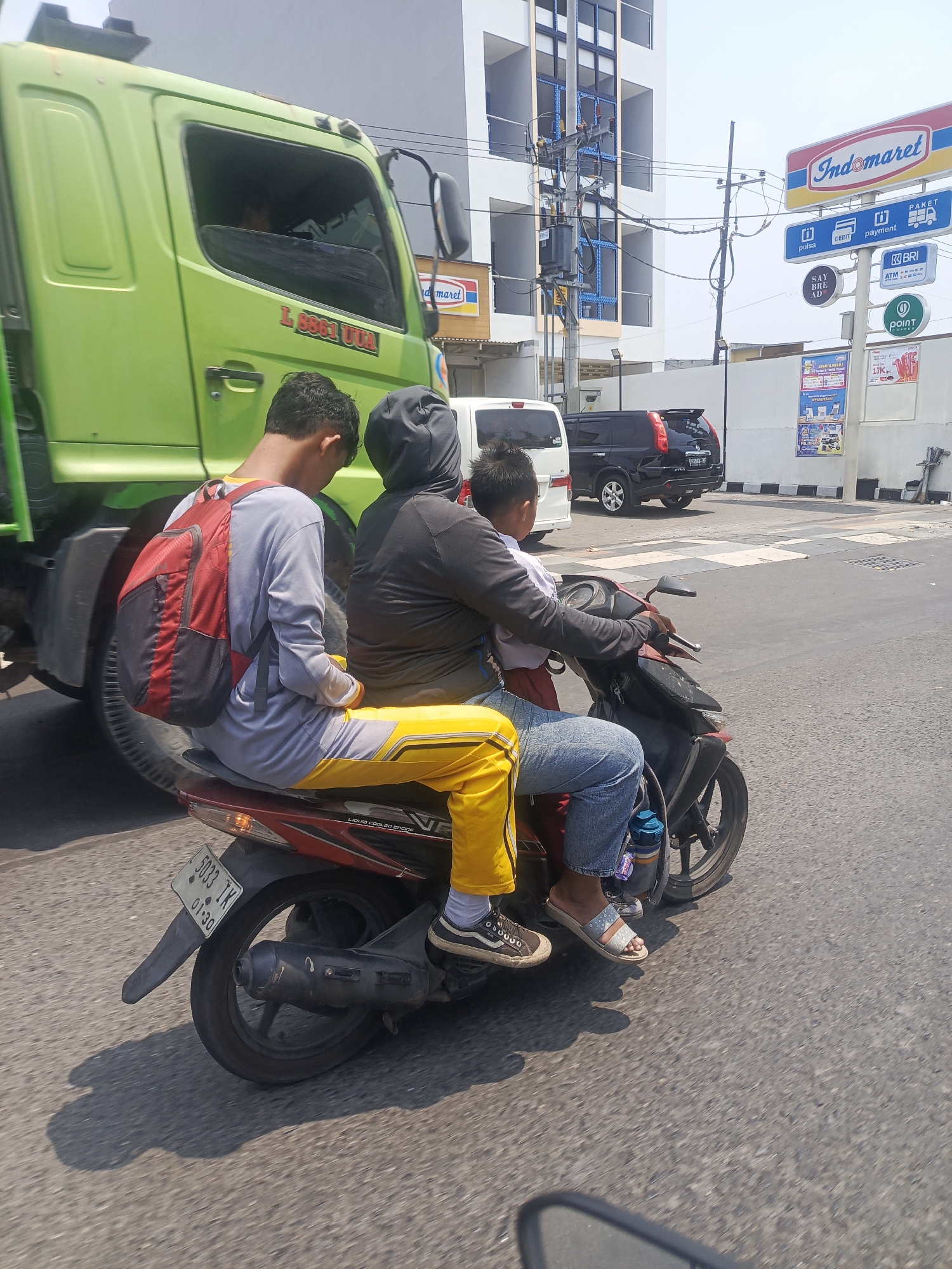 This is a street-level, medium-shot, eye-level photograph taken in Surabaya, Indonesia. In the foreground, three individuals are riding a red motorcycle. The person in the front is wearing a black hooded jacket and blue jeans. Seated behind them is a younger child in a white shirt. The person at the very back is wearing a gray long-sleeved shirt, yellow pants with a stripe, and a red and black backpack. They are all traveling on a busy street, indicated by the presence of a large green truck to the left, also in motion. In the background, there are commercial buildings and street signs, including one for "Indomaret," a convenience store chain. The sky is bright and clear, suggesting a sunny day. The overall scene depicts everyday transportation in a commercial area of Surabaya.