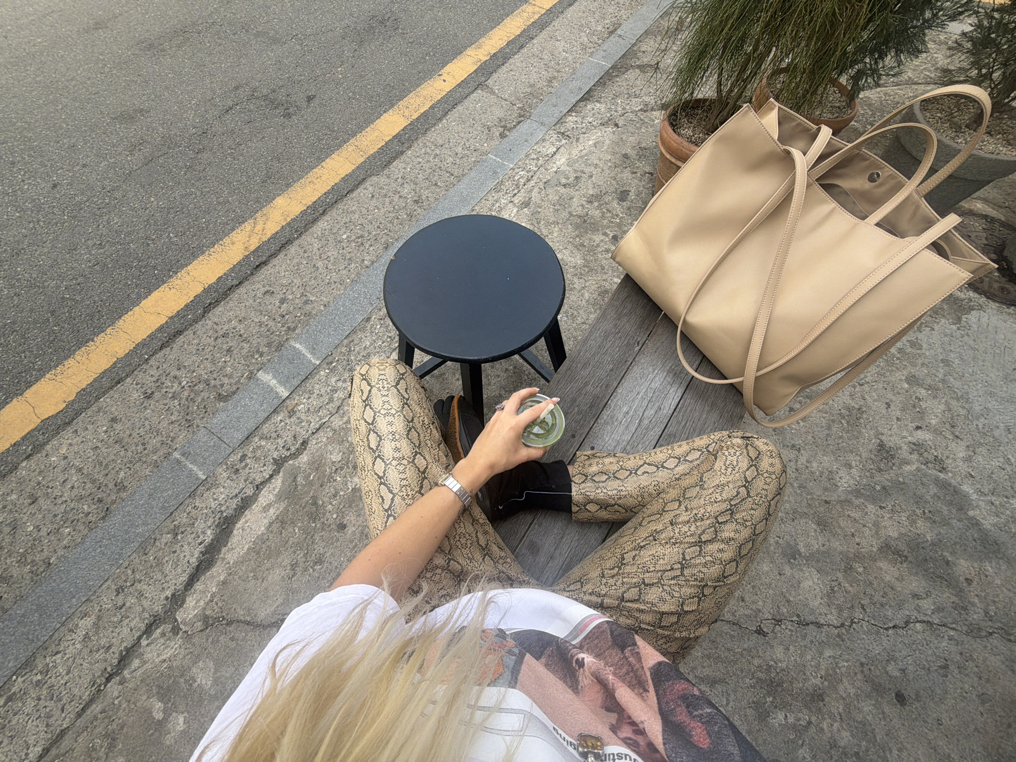 The scene is an overhead shot of a woman sitting at a small, black, round stool on a wooden bench outside in an urban setting, likely Seoul, South Korea.
She is wearing tan, snakeskin-print pants, black shoes, a white t-shirt with an image printed on it, and a silver wristwatch. Her long blonde hair is partially obscuring the view of her shirt. She is holding a green drink in a clear glass with a lid in her right hand.
Beside her, on the right, is a large tan tote bag. Behind the bag, there is a potted plant with green grass-like foliage.
To the left of the woman and the stool is a road with a yellow line marking the edge. The surface the woman is sitting on is a mixture of concrete and wooden planks. The overall atmosphere is casual and relaxed, suggesting a moment of leisure in a city environment.