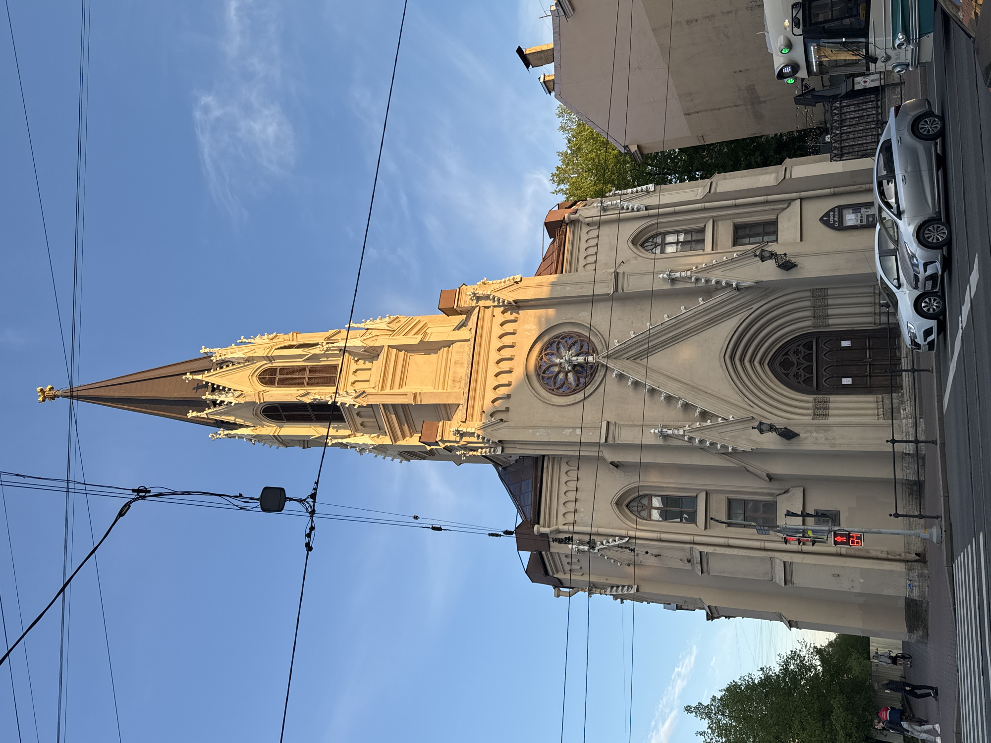 The image shows the facade and steeple of St. Stanislaus Church in Saint Petersburg, Russia, from a slightly low angle, oriented sideways in the frame. The church is made of light-colored stone with Gothic Revival architectural details, including pointed arches, windows with tracery, and a tall, slender steeple. A rose window is visible on the facade.
The sky is blue with wispy clouds. Electrical wires crisscross the upper part of the image. A section of street with a crosswalk is visible, along with a parked silver car and the rear of a bus or trolley. A traffic signal pole shows a red light, and a pedestrian countdown timer displays the number "19." Trees appear in the background at the bottom right, and indistinct figures of people can be seen on the sidewalk.