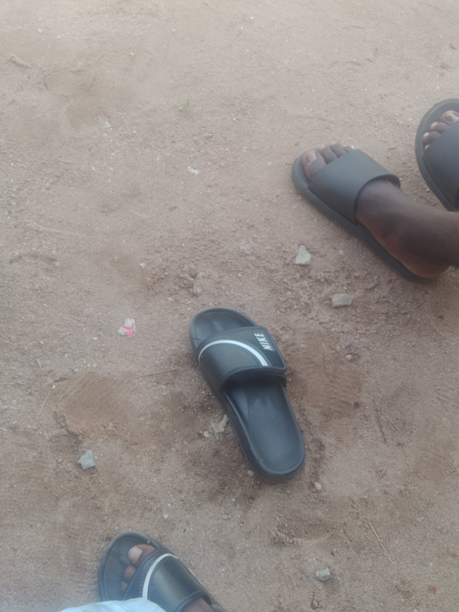 Three pairs of dark-colored slide sandals are visible on a sandy, dirt ground surface in Jauro Yinu, Nigeria. One pair of sandals, with a white stripe across the top, is positioned in the center of the frame and displays the word "NIKE" on the strap. Another pair of sandals is visible on the right side of the frame, with a foot wearing one of them. A third pair of sandals is partially visible at the bottom of the frame, with a foot wearing one of them. The ground is a mixture of sand and small pebbles, with some faint tire tracks and scattered debris, including a small piece of red and white paper. The lighting suggests it is daytime.