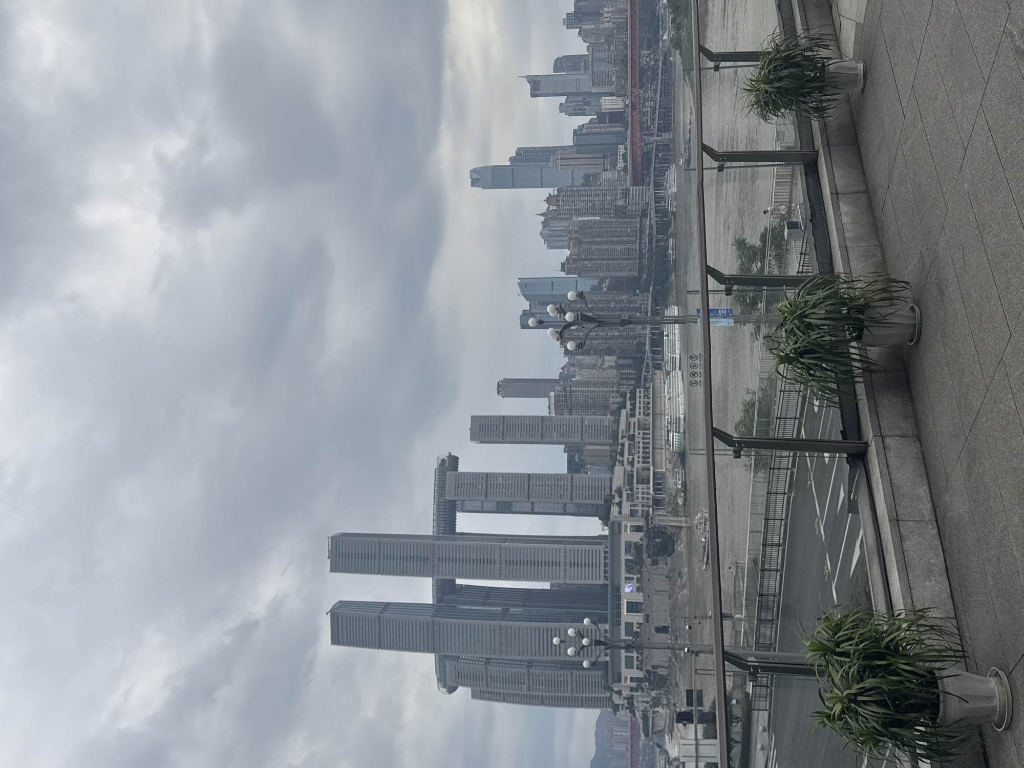The image shows a skyline of a city in Nan'an District, China, taken from an elevated observation point. The buildings are a mix of high-rise structures, with some showing distinctive architectural designs, including towers that appear to be linked by sky bridges. The weather is overcast, with heavy clouds dominating the sky. The foreground features a gray tiled surface with potted plants along the edge, and a glass barrier separates the viewing area from the urban landscape below. A river or large body of water is visible between the observation point and the buildings. There are no people visible in the scene.