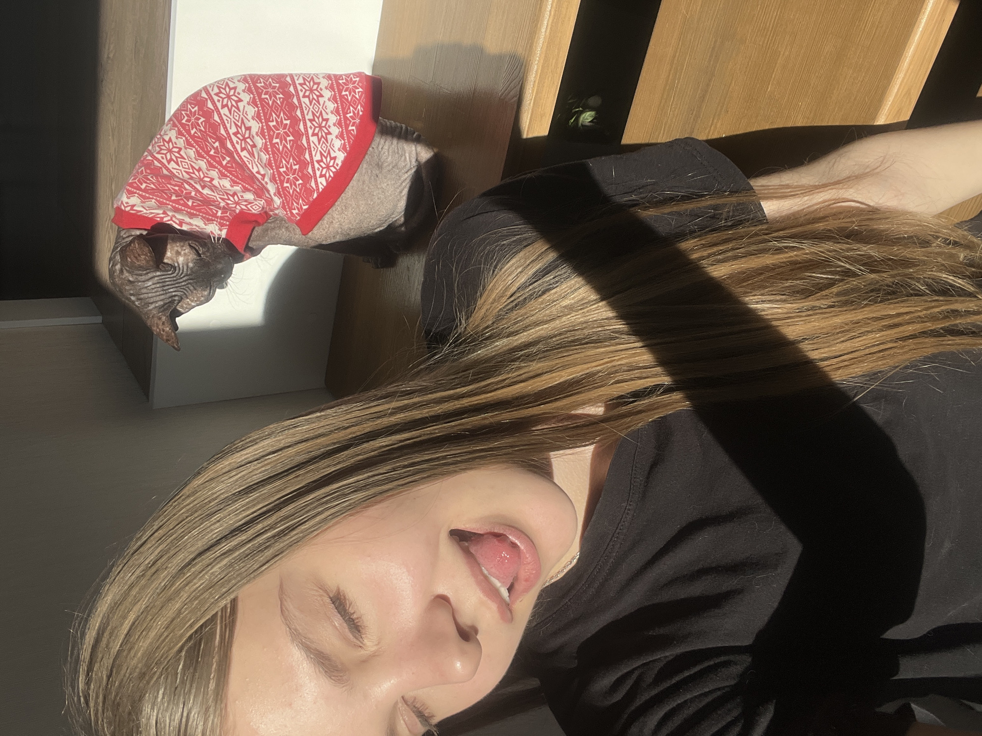 A woman with long, blonde hair is lying down, her face close to the camera. She has her eyes closed and is sticking her tongue out. She is wearing a black shirt. Behind her is a hairless cat wearing a red and white sweater. They appear to be indoors, near a light-colored wall and a wooden structure. The image does not provide information to ascertain the location as Votkinsk, Russia.