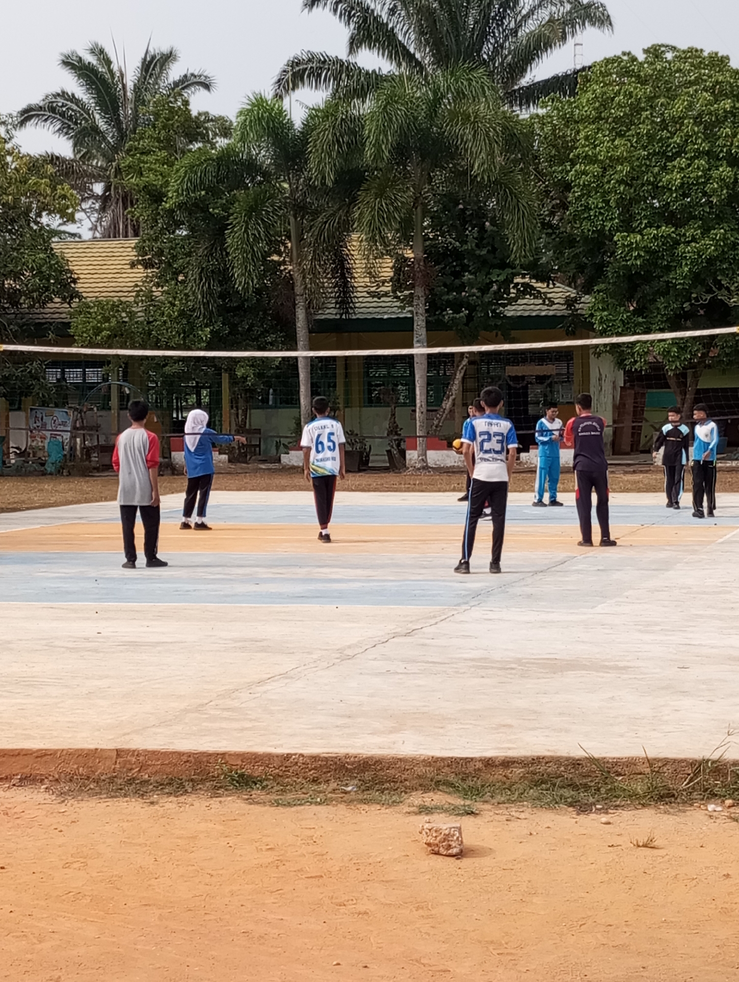 A group of young people, seemingly students, are engaged in a volleyball game on an outdoor court in Pematang Reba, Indonesia. The court is marked with distinct lines in blue, white, and orange. A white net divides the court, with players positioned on both sides. One player in the foreground on the left wears a red and gray long-sleeved shirt and black pants. On the right side of the court, a player in a white shirt with blue accents and the number "65" on the back is visible. Further back, another player wears a blue shirt with the number "23" and black pants. Several other individuals are scattered across the court, some observing and others participating. The background features a building with a yellow roof and palm trees, suggesting a school or community setting. The ground surrounding the court is a mix of dirt and concrete. The sky is overcast.