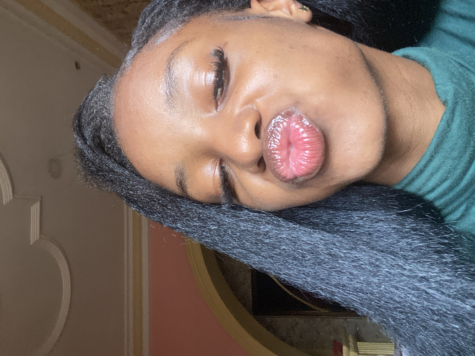The image is a close-up portrait of a young Black woman, captured indoors in what is identified as Jos, Nigeria. She is positioned centrally, slightly angled to her right, with her head tilted slightly downwards. Her eyes are closed or heavily squinted, and her lips are prominently pursed into a "kissing" expression.
Her skin has a dark complexion, appearing smooth and hydrated, with noticeable shine on her forehead and cheekbones. She has long, dark eyelashes, likely false or enhanced, and natural, well-defined eyebrows. Her pursed lips are coated with a high-shine, glossy, pinkish-red product, showcasing vertical lines. A small, gold-colored, star-shaped earring is visible in her right earlobe. Her hair consists of long, dark, heavily textured extensions or braids, which cascade over her left shoulder, filling a significant portion of the left side of the frame. Visible baby hairs frame her forehead. She is wearing a high-neck, ribbed top in a deep emerald green color.
The background depicts an interior room. The upper section of the wall or ceiling is light beige or off-white, adorned with decorative white molding, including a linear band and a semi-circular architectural motif. Below this, on the left side of the frame, a segment of a peach or light terracotta colored wall is visible. To the right of the subject, a dark-framed archway or doorway is partially visible, offering a glimpse into an adjacent space or feature, which appears to include a small, ornate structure, possibly a mantle, with a reddish object resting on it. The lighting is soft and even, highlighting the subject's facial features.