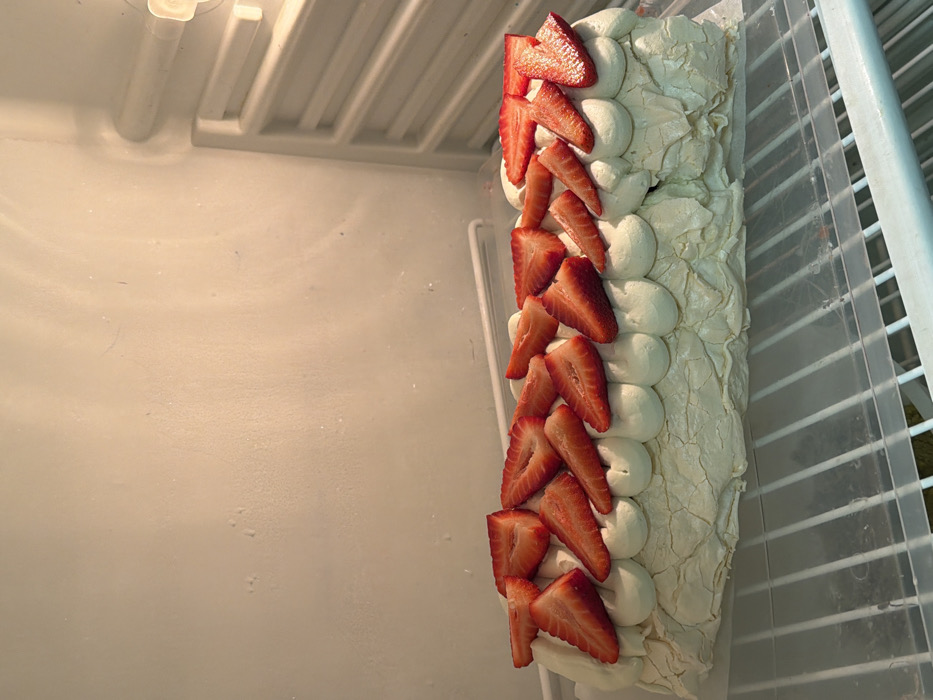 A long, rectangular meringue dessert, likely a pavlova roll, is positioned horizontally on a clear, plastic-covered wire shelf inside a refrigerator. The dessert features a textured, pale off-white meringue base with visible cracks. Its top is adorned with numerous mounds of piped, light-colored cream, upon which multiple freshly sliced strawberries are arranged. The strawberries are cut lengthwise into halves or quarters, revealing their red interior flesh and some lighter pith. The interior of the refrigerator is stark white, with textured walls and vertical shelving supports visible on the right side. A light source is located in the upper right corner, illuminating the appliance's interior. No specific city location within Tel-Aviv, Israel, can be determined from the visual evidence provided. No people are visible.