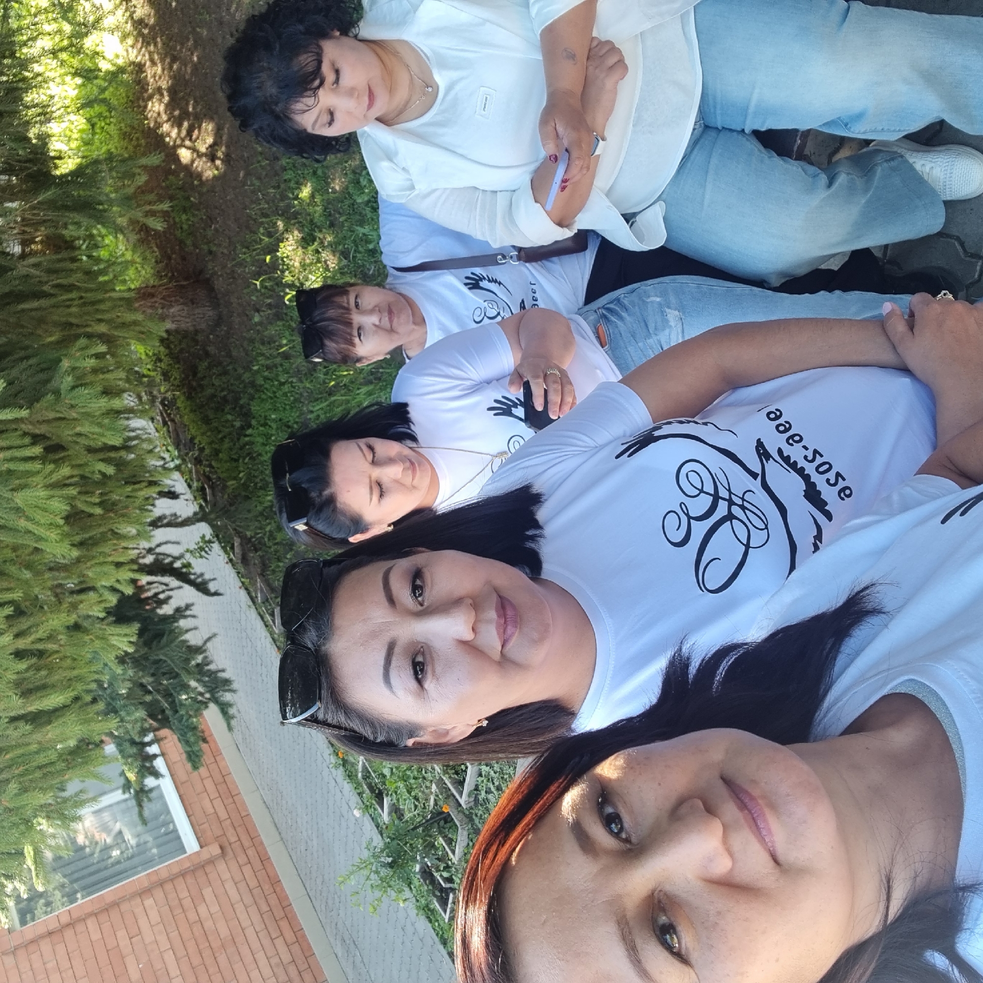 In the city of Aktobe, Kazakhstan, five women are pictured outdoors, seated on a bench under a large coniferous tree. The setting appears to be a public space with a brick building and paved walkway visible in the background. The women are dressed casually in white t-shirts and denim jeans. Three of the women are prominently featured in the foreground, with the other two visible behind them. The woman on the far left is looking directly at the camera. The woman next to her is also looking forward, while the woman in the middle is looking down at her phone. The woman to her right is looking forward, and the woman on the far right is looking down at her phone. The image is taken during the day, with sunlight filtering through the trees, creating dappled shadows on the ground.