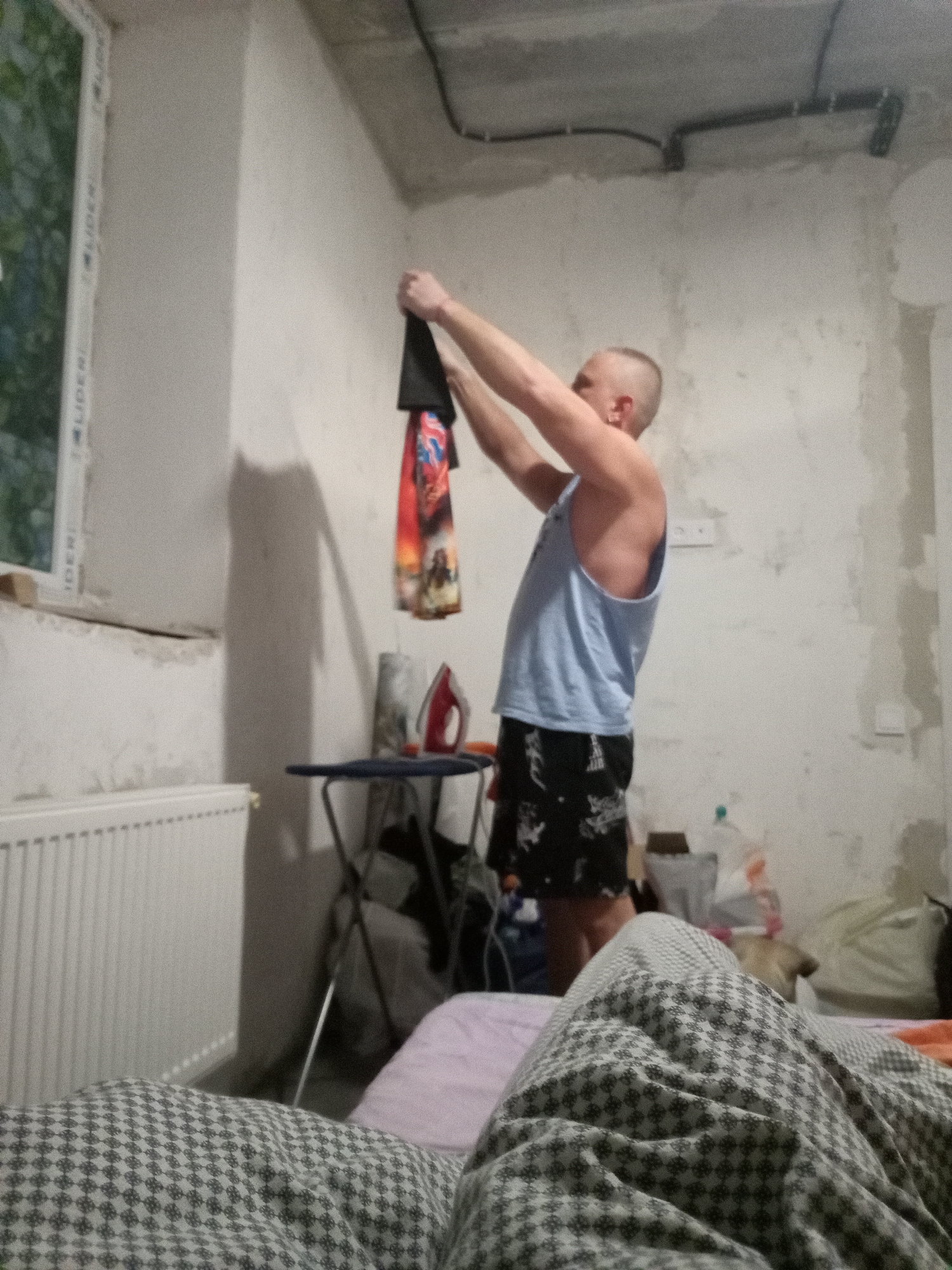 The image depicts an indoor scene, likely a bedroom or unfinished room, in an unknown location. A person is standing in the center of the frame, holding up a black garment with a colorful design, possibly a shirt or flag. They are wearing a light blue tank top and black shorts with a white pattern. To the left of the person, a window with green foliage visible outside is present, and below it, a white radiator runs along the wall. In the mid-ground, an ironing board with an iron on it is set up. The walls are rough and unfinished, with exposed electrical conduits visible on the ceiling. The foreground is occupied by a patterned blanket, suggesting a bed. The lighting is dim, with a strong shadow cast on the wall behind the person.