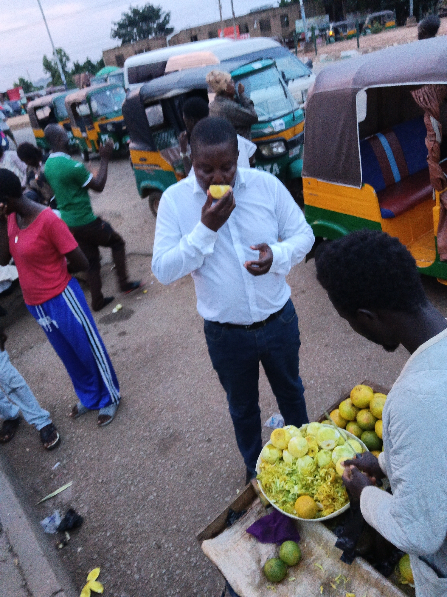 In Bukuru, Nigeria, on a dirt road with some scattered debris, a man in a white long-sleeved shirt and blue jeans is eating a piece of yellow fruit, possibly a lemon or lime, held in his right hand. He is standing next to a cart laden with a large quantity of green and yellow citrus fruits, some of which are peeled, and shredded green rind. To the right of the fruit cart, another man, seen from the shoulders down, is wearing a light-colored garment and appears to be preparing fruit. In the background, several colorful auto-rickshaws are parked. Other individuals, partially visible, are also present in the scene. The lighting suggests it is late afternoon or early evening.