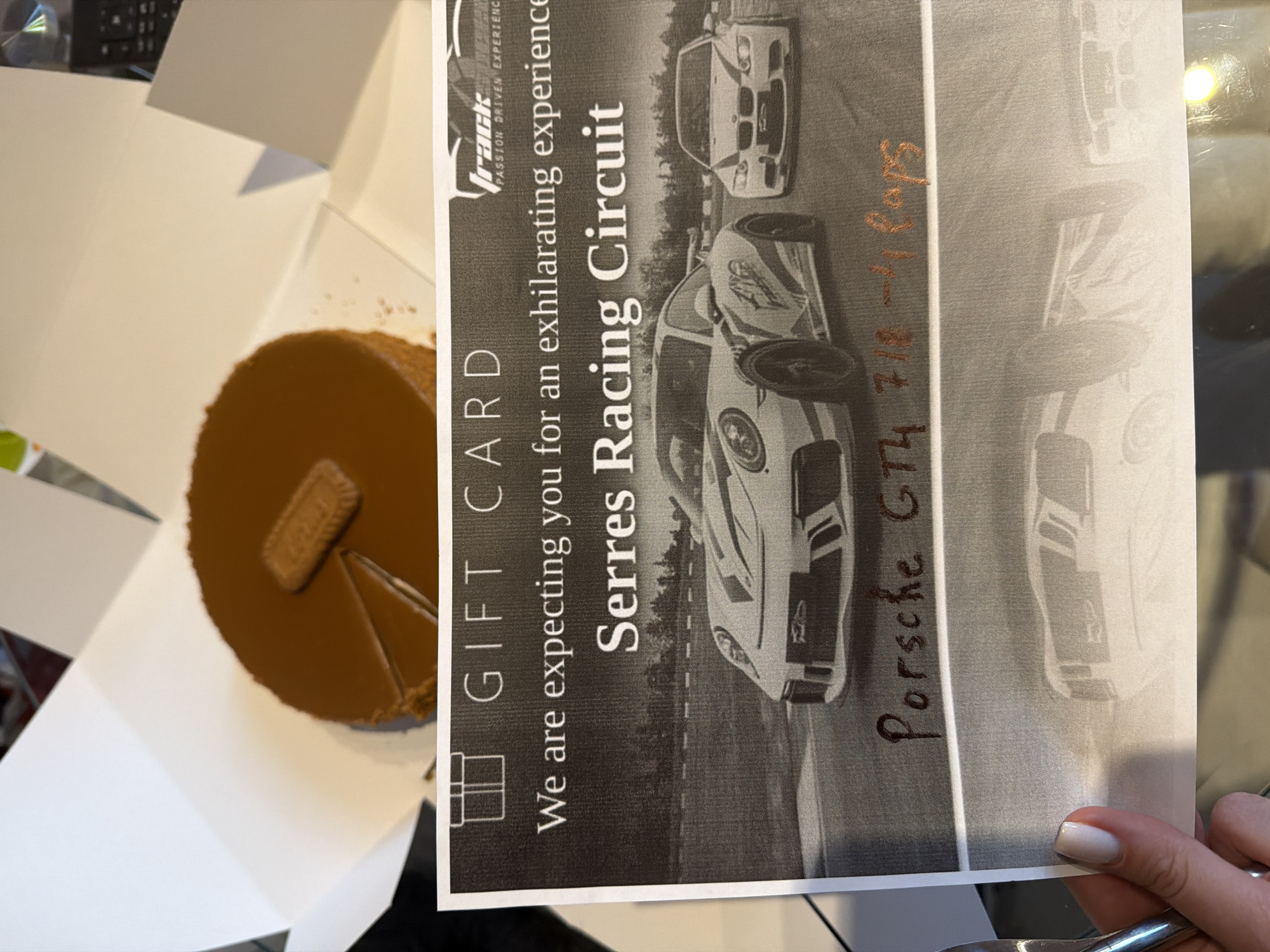 The image shows a gift card for "Serres Racing Circuit" with an image of sports cars on a track. The gift card is partially obscured by a cake with a Lotus biscuit on top, placed in a white box. Below the cars, handwritten text in dark red reads "Porsche GT4 718-4 laps". The scene is indoors, with a table surface visible below the gift card. There is no visible connection to the city of Sofia, Bulgaria.
