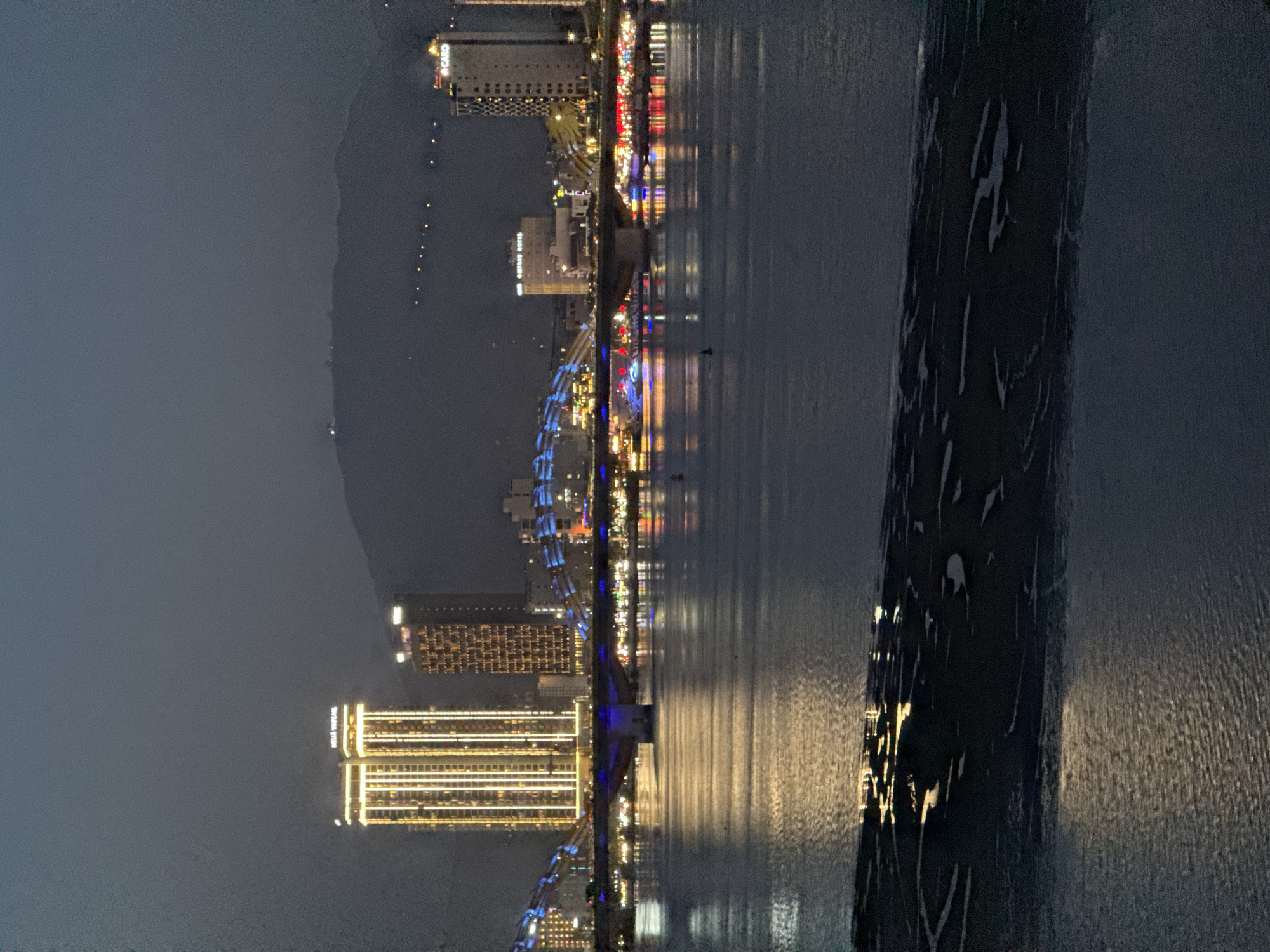 This is a nighttime photograph taken from the shore of the Han River in Da Nang, Vietnam, looking towards the city skyline. The water in the foreground reflects the lights from the city and the bridge. The iconic Dragon Bridge is visible spanning the river, illuminated with blue lights. On the far bank, modern skyscrapers, including the Novotel Da Nang Premier Han River and the Muong Thanh Grand Da Nang Hotel, are lit up. The background features a dimly lit mountain range. The overall atmosphere is one of urban nighttime vibrancy.