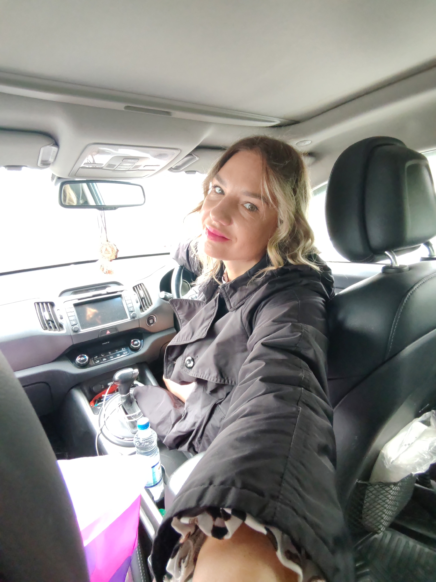 A woman is seated inside a car, facing the camera. She is wearing a dark-colored jacket. The car's interior is visible, including the dashboard, steering wheel, and gear shift. A water bottle and a shopping bag are in the center console. The location is presumed to be in the city of Petropavlovsk-Kamchatsky, Russia.