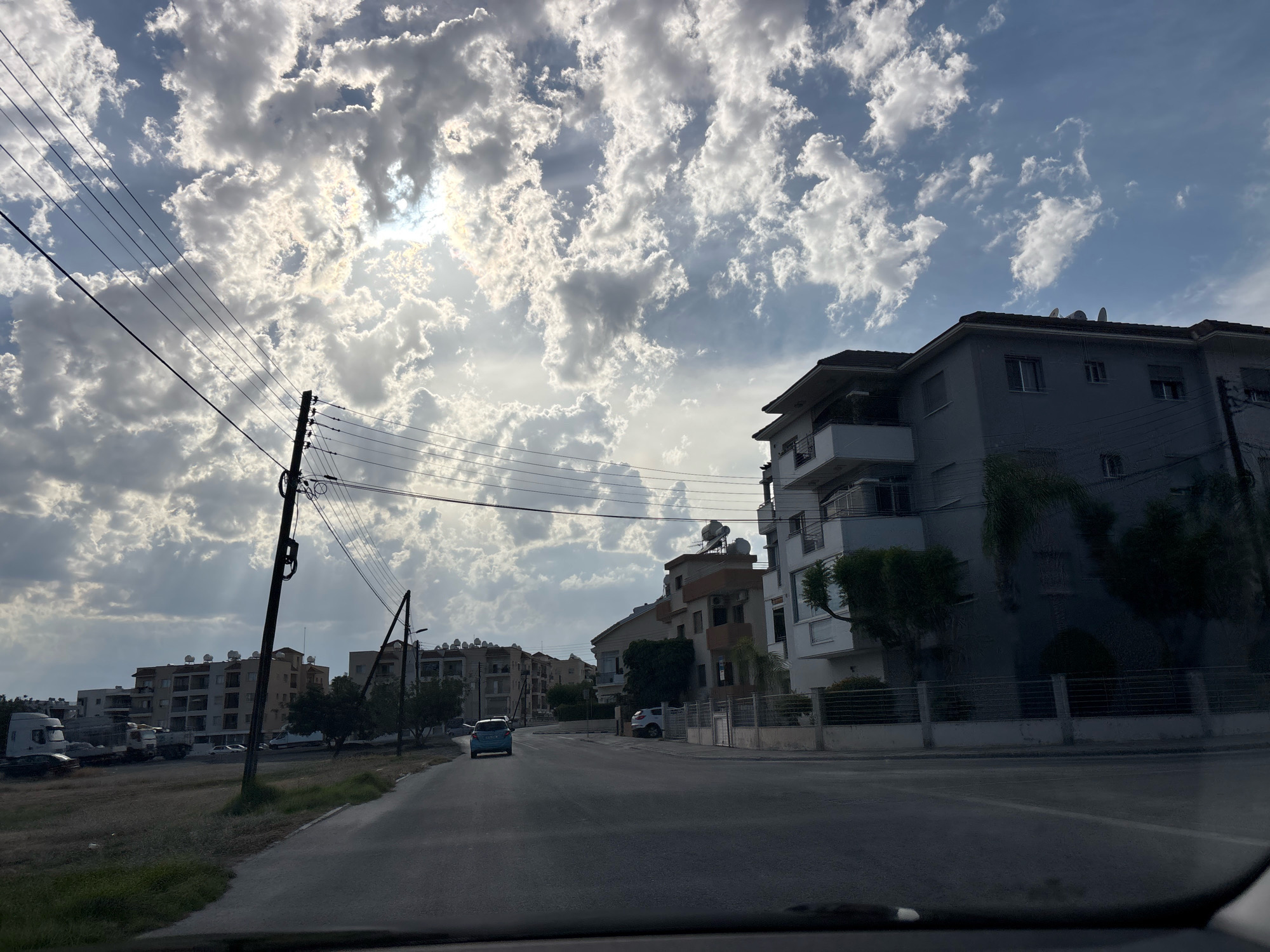 The image depicts a street scene in Agios Athanasios Municipality, Cyprus. The sky is filled with dramatic, cloud-filled skies with sunlight peeking through. Power lines and utility poles are prominent in the foreground and mid-ground. On the right side of the street, a multi-story residential building with balconies is visible, featuring a light-colored facade and a dark roof. Adjacent to it, a similar building with a darker facade can be seen. Further down the street, other apartment buildings with multiple floors and visible satellite dishes are present. To the left of the street, an open field with dry vegetation is visible, with several construction sites or unfinished buildings and parked trucks. A light blue car is driving away from the camera on the asphalt road, and a white car is parked on the right side of the road. Trees are planted along the street and in front of the buildings. The overall impression is of a developing urban or suburban area under a cloudy sky.