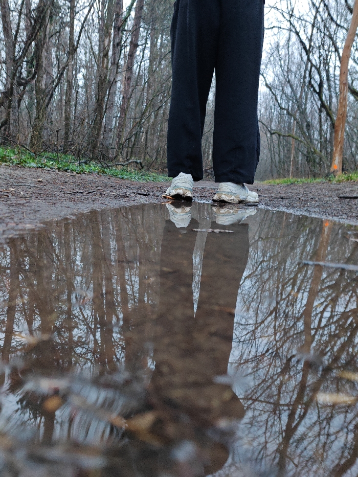 The scene depicts the lower extremities of an individual standing on a dirt trail in a deciduous forest, viewed from a low perspective. A large, stagnant puddle of muddy water occupies the foreground, reflecting the subject and the surrounding environment.
**Location:** This scene is located on a forest trail, likely within the Shumen Plateau Nature Park, Shumen, Bulgaria.
**Subject:** A person stands with their back partially to the camera. They are wearing dark-colored, loose-fitting trousers and light-colored athletic shoes. The shoes show evidence of dirt or mud on their uppers and soles. The individual's legs and shoes are distinctly reflected in the puddle's surface.
**Environment:** The background consists of a dense forest composed primarily of tall, bare deciduous trees, suggesting late autumn or winter. The ground is a dirt path, with sparse green vegetation visible along its edges, particularly to the left. The sky, partially visible through the tree canopy, is overcast and light grey. A tree trunk on the right side of the frame exhibits an orange marking, indicative of a trail marker. The puddle itself contains sediment and reflects the overhead trees and sky with clarity. The ground adjacent to the puddle appears damp.