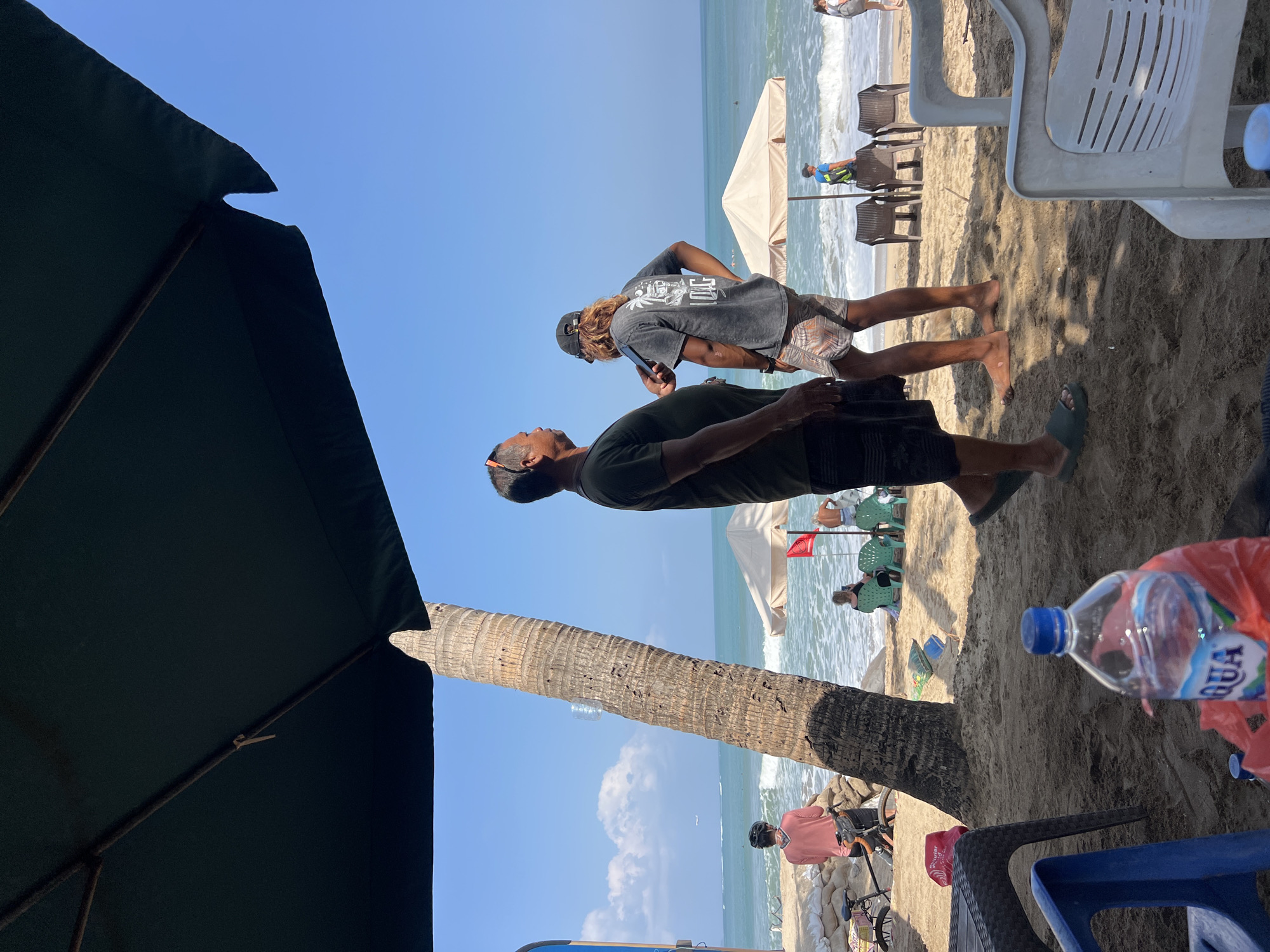 On a sunny day at Kuta Beach, Indonesia, two men stand on the sand under a large dark umbrella. The man on the left, wearing a dark t-shirt and shorts, faces away from the camera. The man on the right, with long wavy hair and wearing a graphic t-shirt and patterned shorts, looks towards the left and appears to be holding a phone.
In the background, the ocean displays gentle waves under a clear blue sky with scattered clouds. A palm tree trunk is prominent on the left side of the frame, casting a shadow. Several beach umbrellas, lounge chairs, and people relaxing on the beach are visible further down the shoreline. A person in a pink shirt on a bicycle is positioned near sandbags on the left side of the image. In the foreground, a plastic water bottle with an "AQUA" label and a red plastic bag rest on the sand, near a blue chair and the edge of a woven table.
