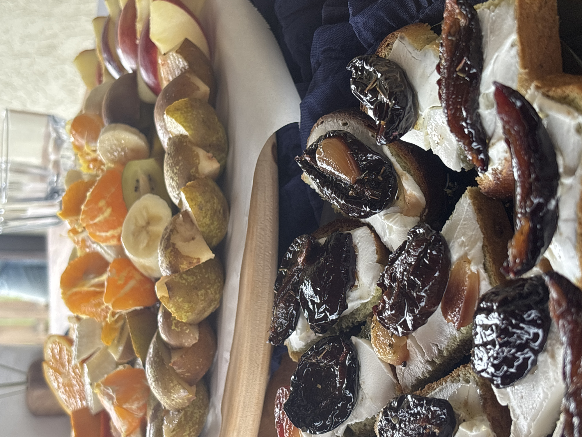 A close-up shot presents a wooden serving tray, likely located in Lahojsk selsaviet, Belarus, containing an assortment of fruit and canapés. On the left side of the tray, arranged in rows, are slices of apple, pear, kiwi, banana, and tangerine. A white, possibly wax paper, divider separates the fruit from the canapés on the right. The canapés consist of small, square pieces of bread topped with a white spread, possibly cream cheese, and dark-colored, shiny fruit, which might be prunes or plums. A dark blue, crumpled cloth is visible under the canapés, providing contrast to the food. The lighting suggests an indoor environment, and the depth of field is shallow, focusing primarily on the food items. No people are directly visible in the frame.