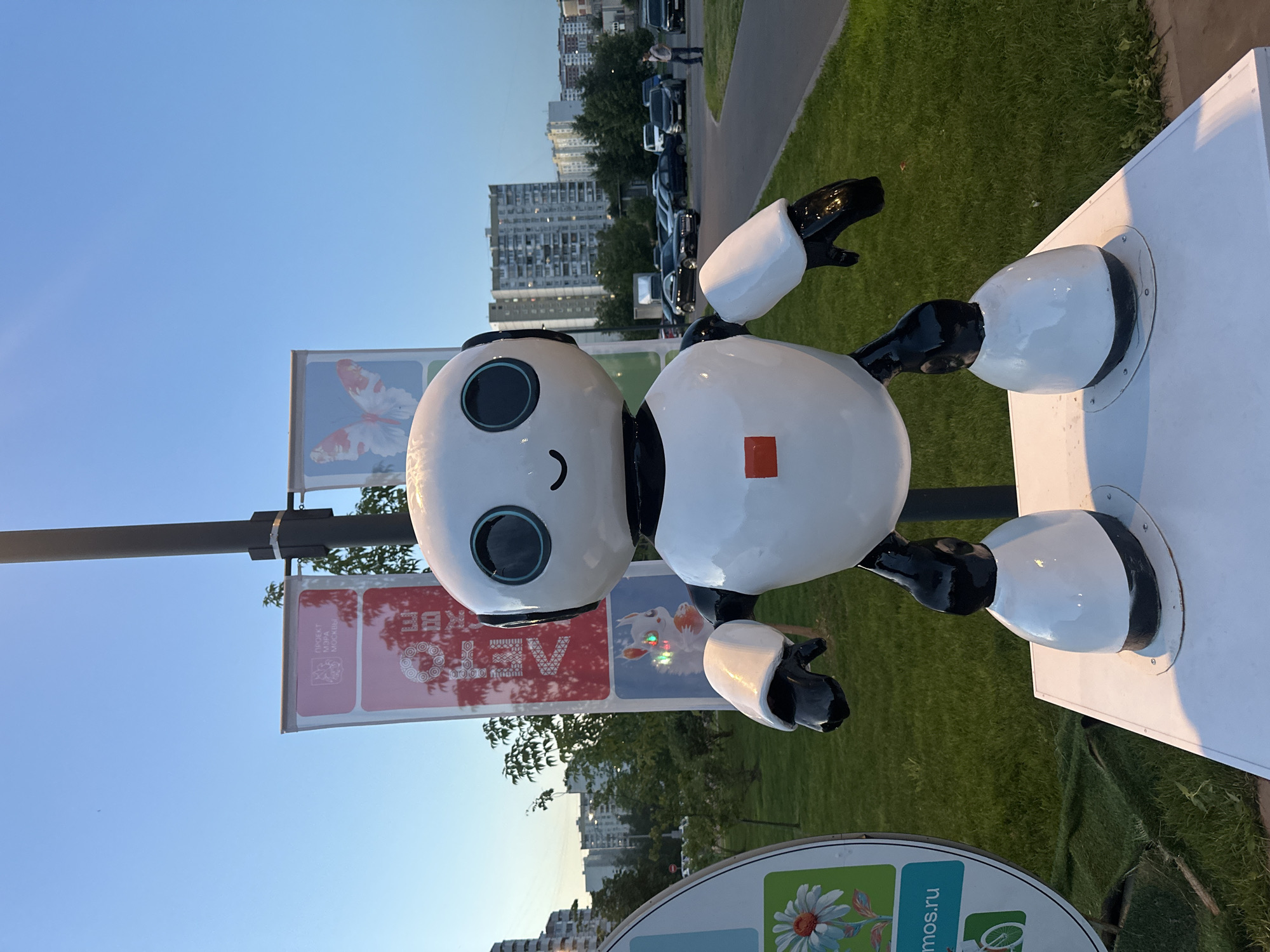 A white robot statue with black limbs and a simple smiley face stands in a park. The robot is positioned on a white pedestal, and behind it, a lamppost holds up two banners. The left banner is red with text and a logo, and the right banner features a white and red butterfly image. In the background, a grassy area leads to a street with several parked cars and residential buildings. The sky is clear and blue. The scene appears to be in Moscow, Russia, as suggested by the text on the banner, which includes "ПРОЕКТ МЭРА МОСКВЫ" (Project of the Mayor of Moscow).