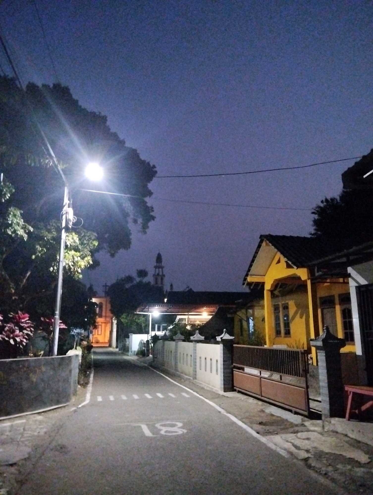 The image depicts a street scene in Salatiga, Indonesia, at dusk or early night. A street lamp on the left illuminates the asphalt road, which has white markings indicating a speed bump or crossing. The number "18" is painted on the road. On the left side of the road, there is a dark wall and some lush green plants. On the right side, there is a yellow house with a dark roof and windows, and a brown metal gate. Further down the street, a building with a tower, possibly a mosque or a church, is visible against the twilight sky. The sky is a deep, dusky blue. Power lines are visible across the sky. The environment appears to be a residential area.